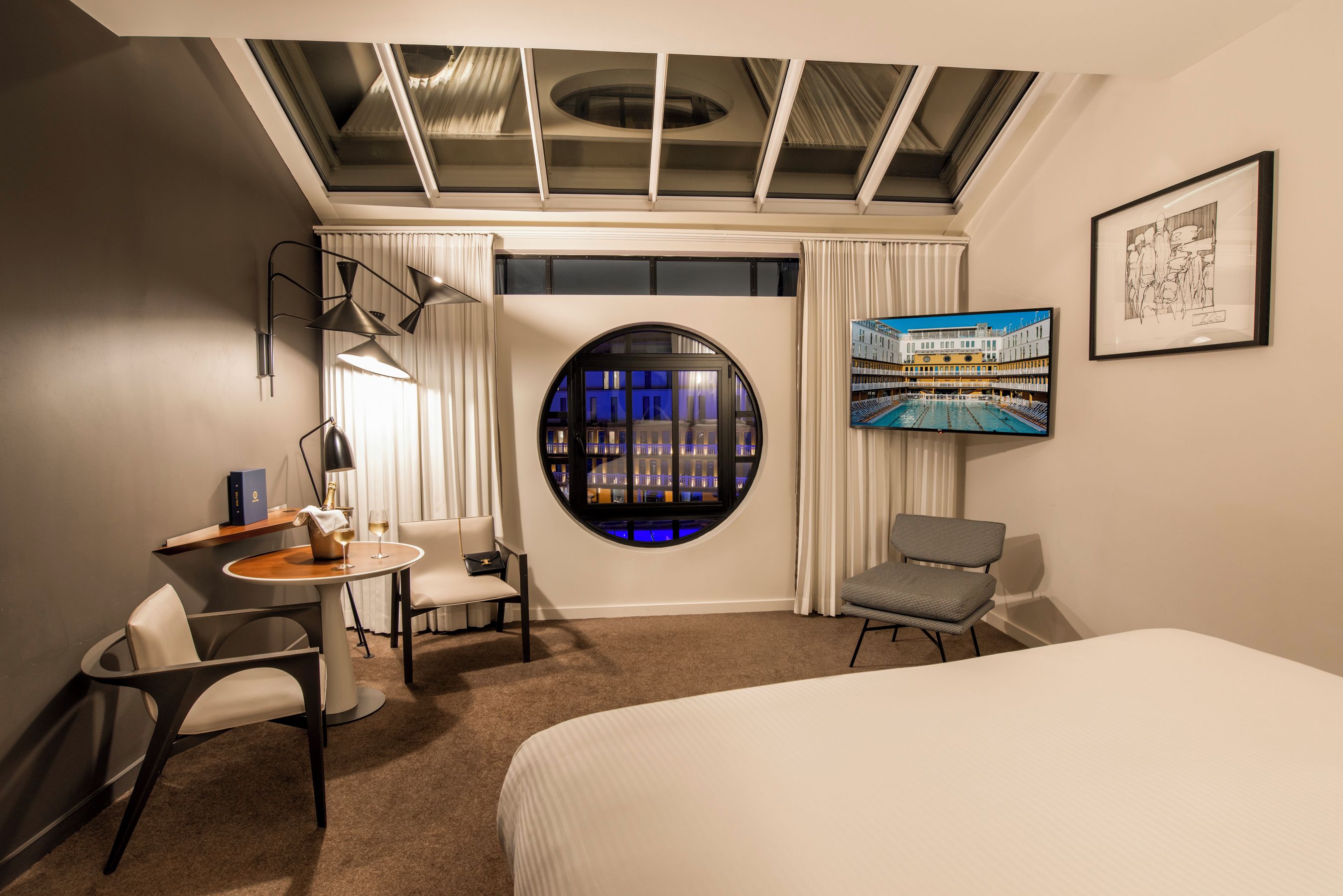 A hotel room with a bed, a desk with two chairs, a round window with a view of buildings, and a flat-screen TV on the wall.