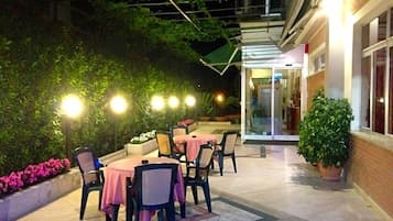 Une terrasse extérieure avec des tables et des chaises, entourée de verdure et donnant sur l’entrée d’un bâtiment.