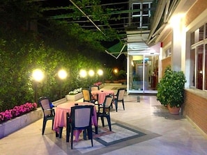 An outdoor patio with tables and chairs, surrounded by greenery and a building entrance.
