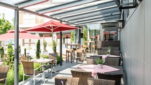 An outdoor patio with wicker furniture, red umbrellas, and glass walls overlooking a street. - Hotel Schiller (Olching)