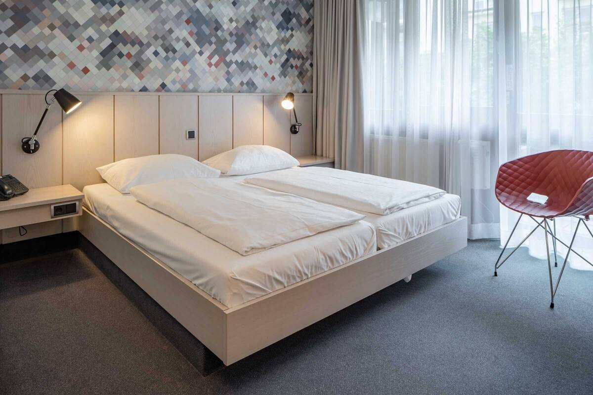 A hotel room with a large bed, two bedside lamps, a red chair, and a patterned wall.