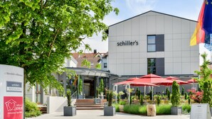 A modern building with a sign reading "schiller's" and outdoor seating with red umbrellas. - Hotel Schiller (Olching)
