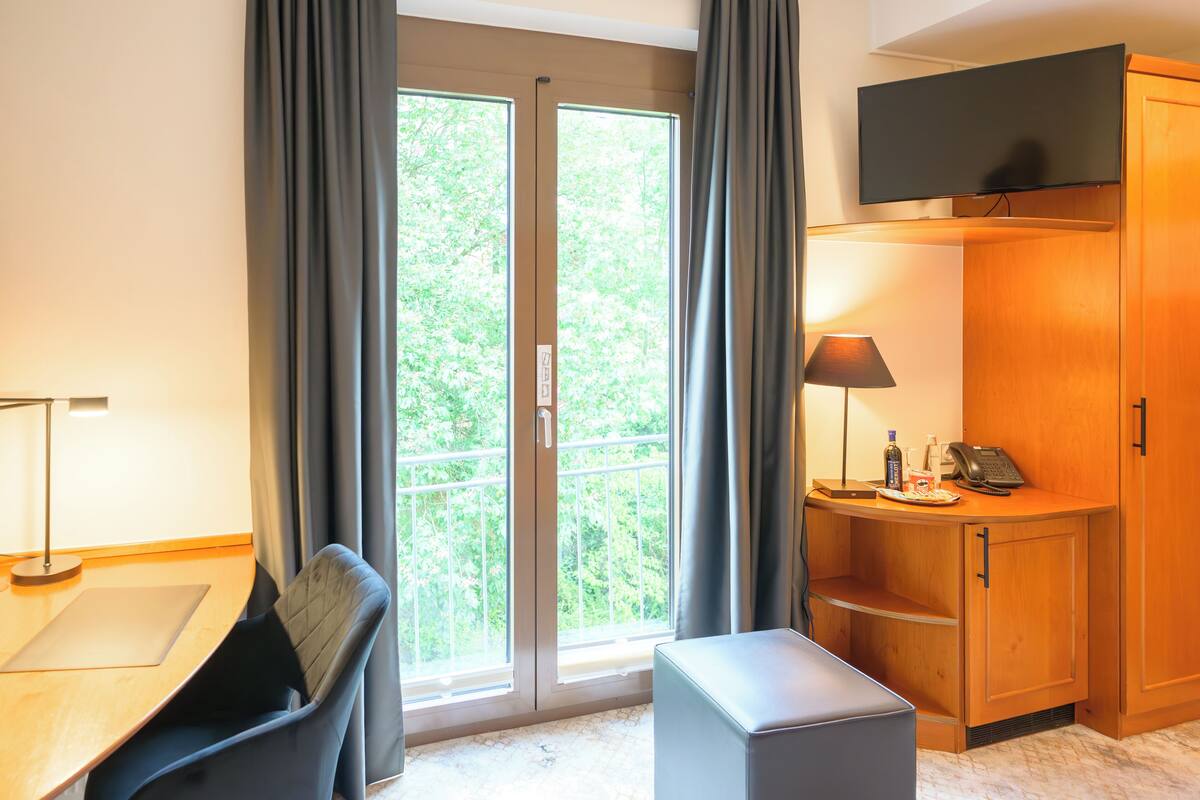 A hotel room with a large sliding glass door to a balcony, a desk with a chair, a small table, a lamp, a cabinet, and a television mounted on the wall.