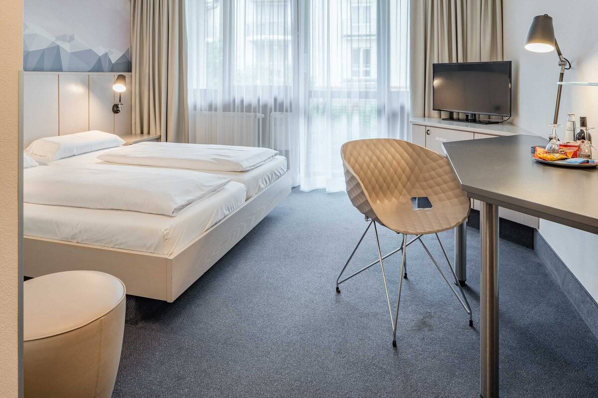 A hotel room with a bed, a desk with a lamp, a chair, and a TV.