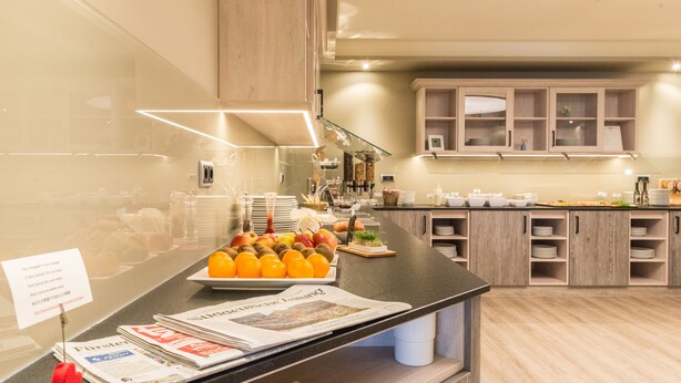 A breakfast buffet area with a variety of fruits and newspapers on the counter. - Hotel Schiller (Olching)
