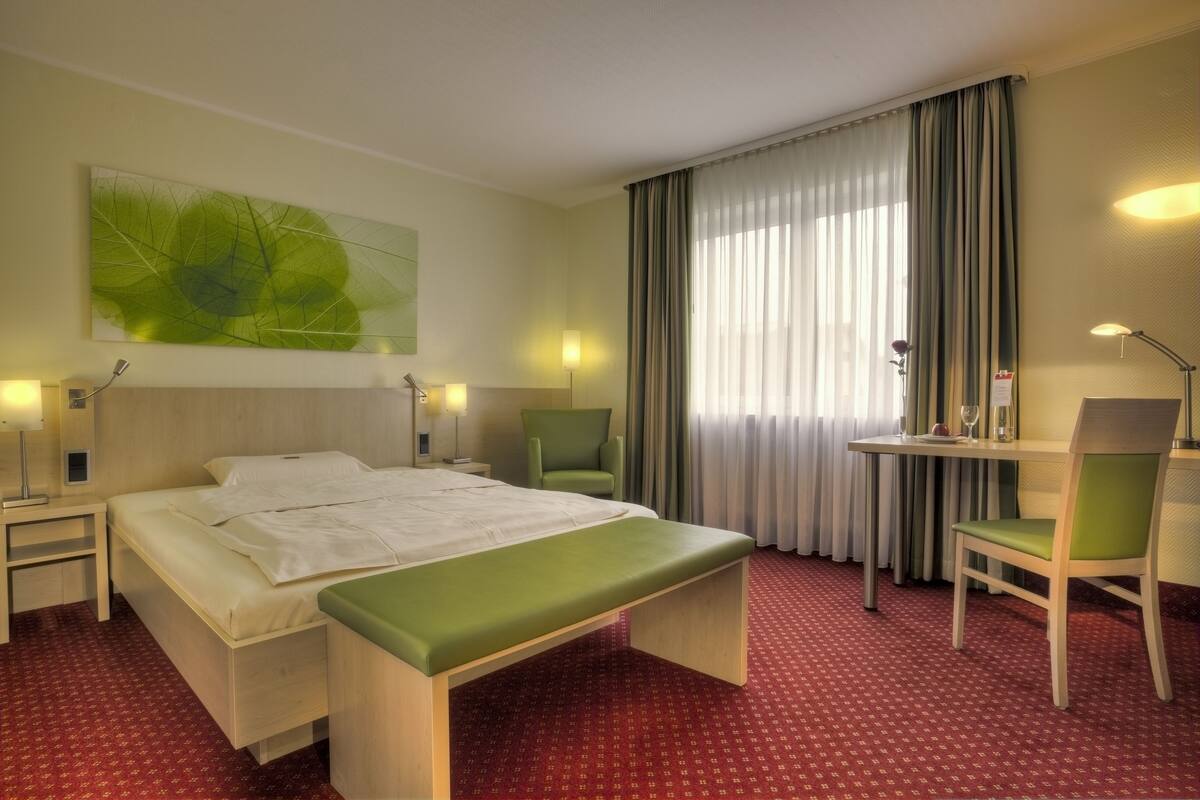 A hotel room with a bed, a desk, a chair, a bench, and a large window with curtains.