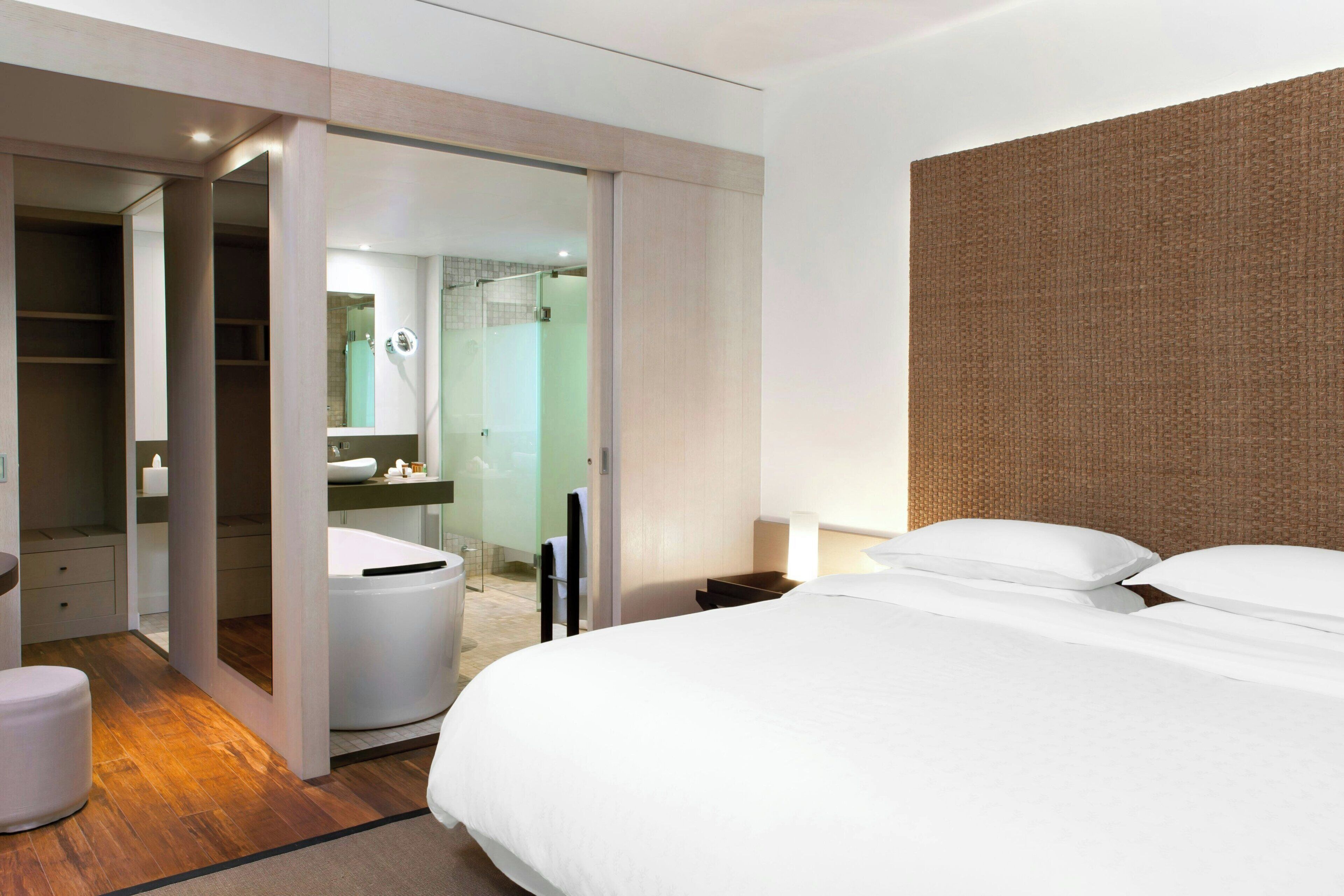 A modern hotel room with a large bed, a walk-in closet, a bathroom with a glass shower, and a wooden headboard.