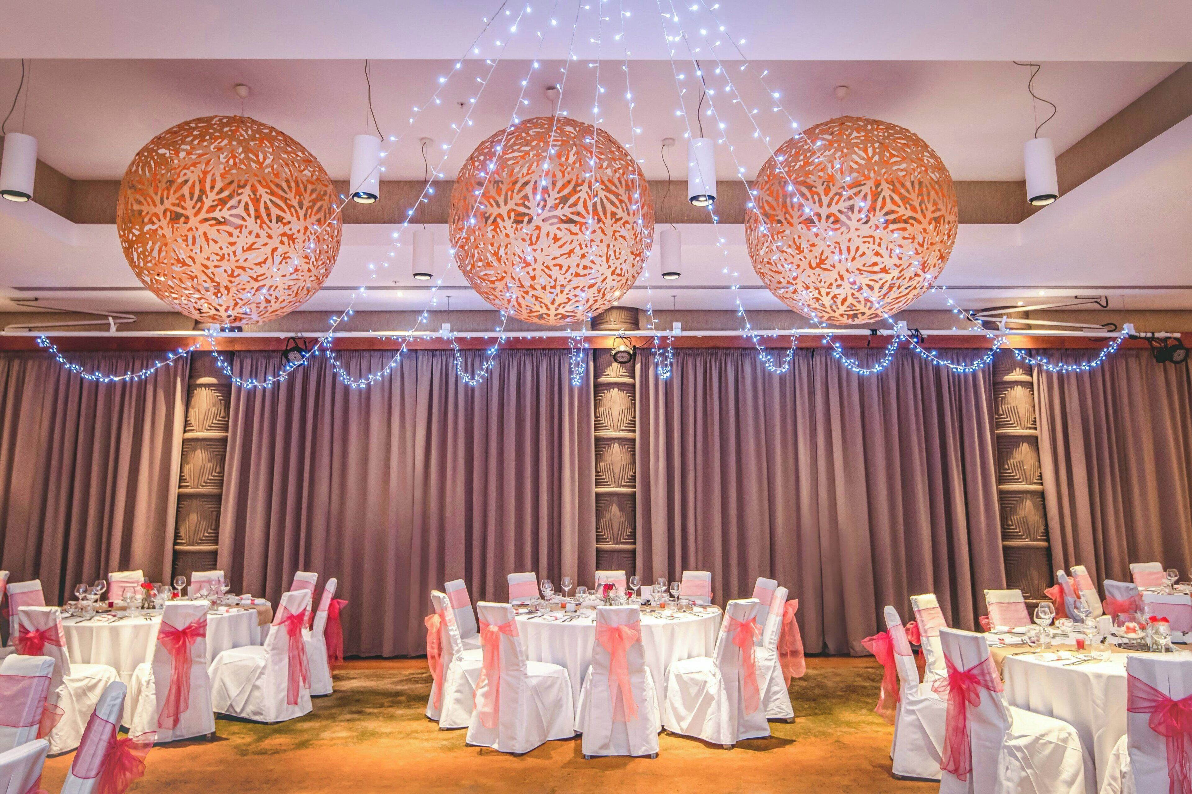Un salón de banquetes con mesas redondas, sillas blancas con cintas rosas e iluminación decorativa colgante.