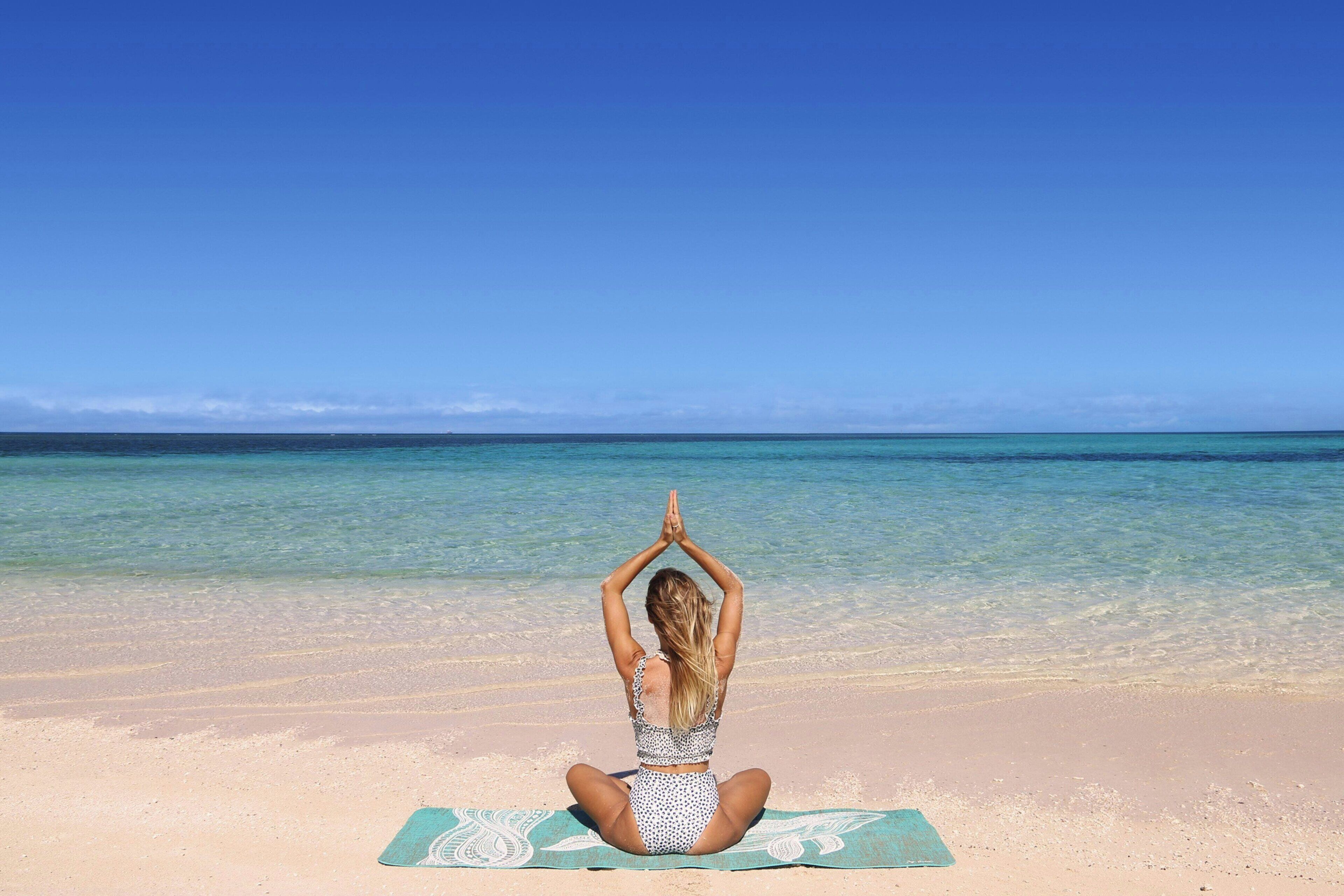 A person sitting on a yoga mat on a beach with clear water and a clear sky.