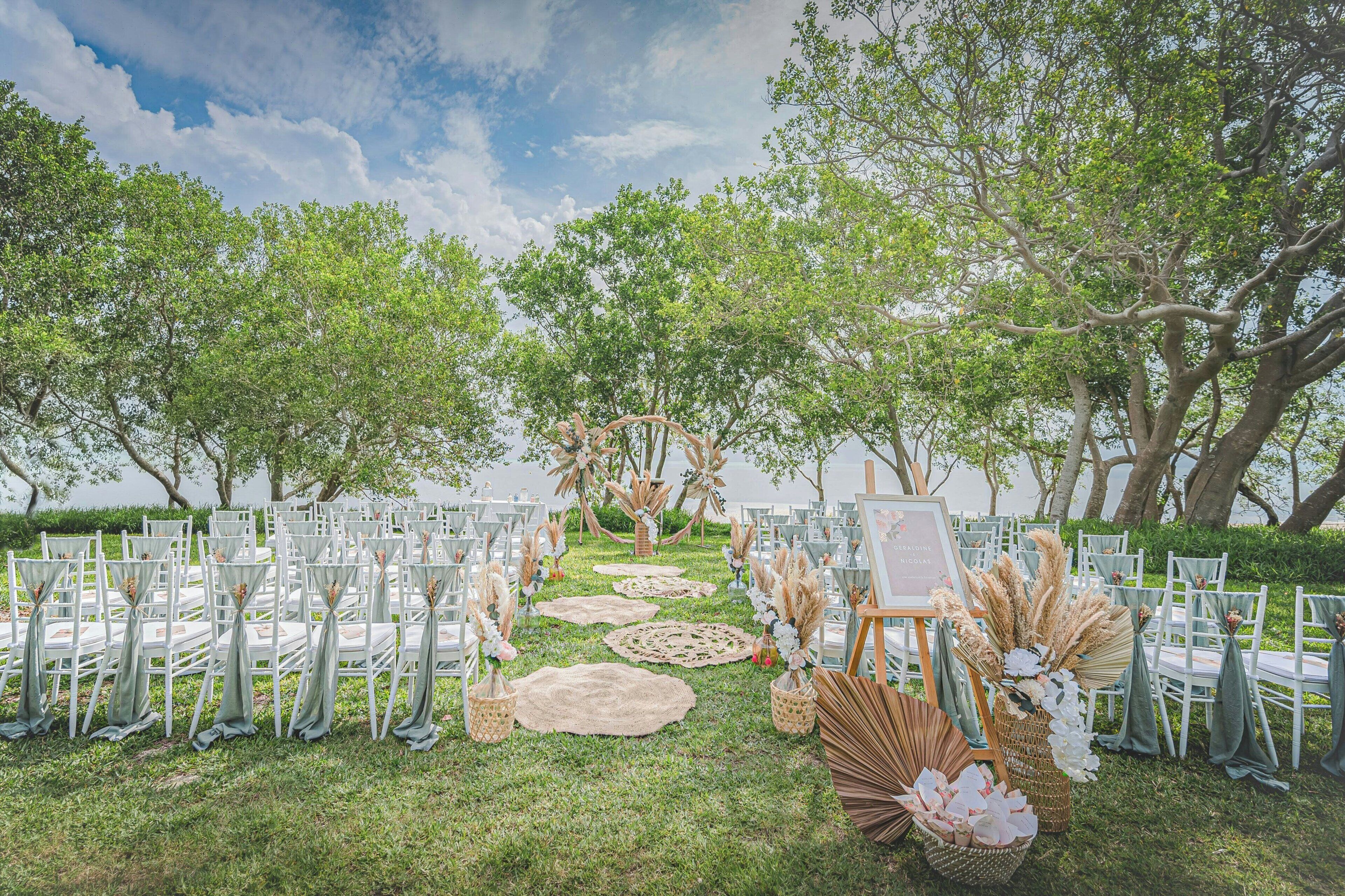 An outdoor wedding venue with rows of white chairs, a grassy aisle, and decorative elements like pampas grass and woven baskets.