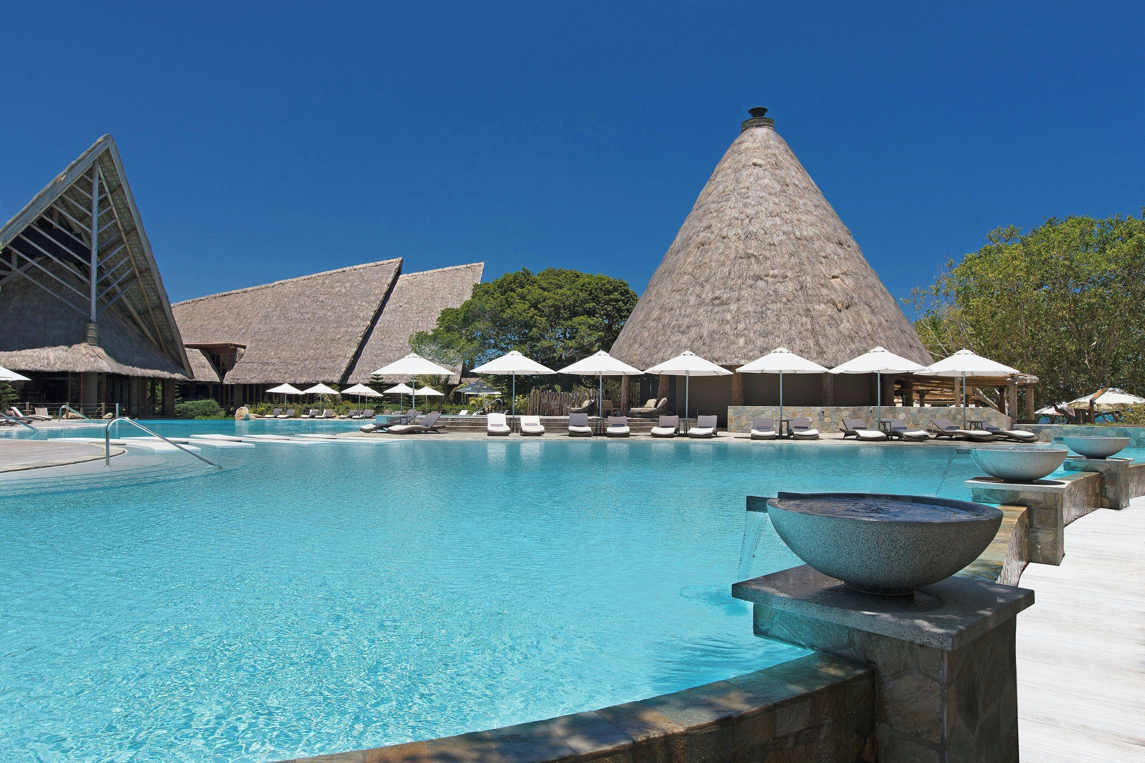 A swimming pool with lounge chairs and umbrellas, surrounded by buildings with conical roofs.