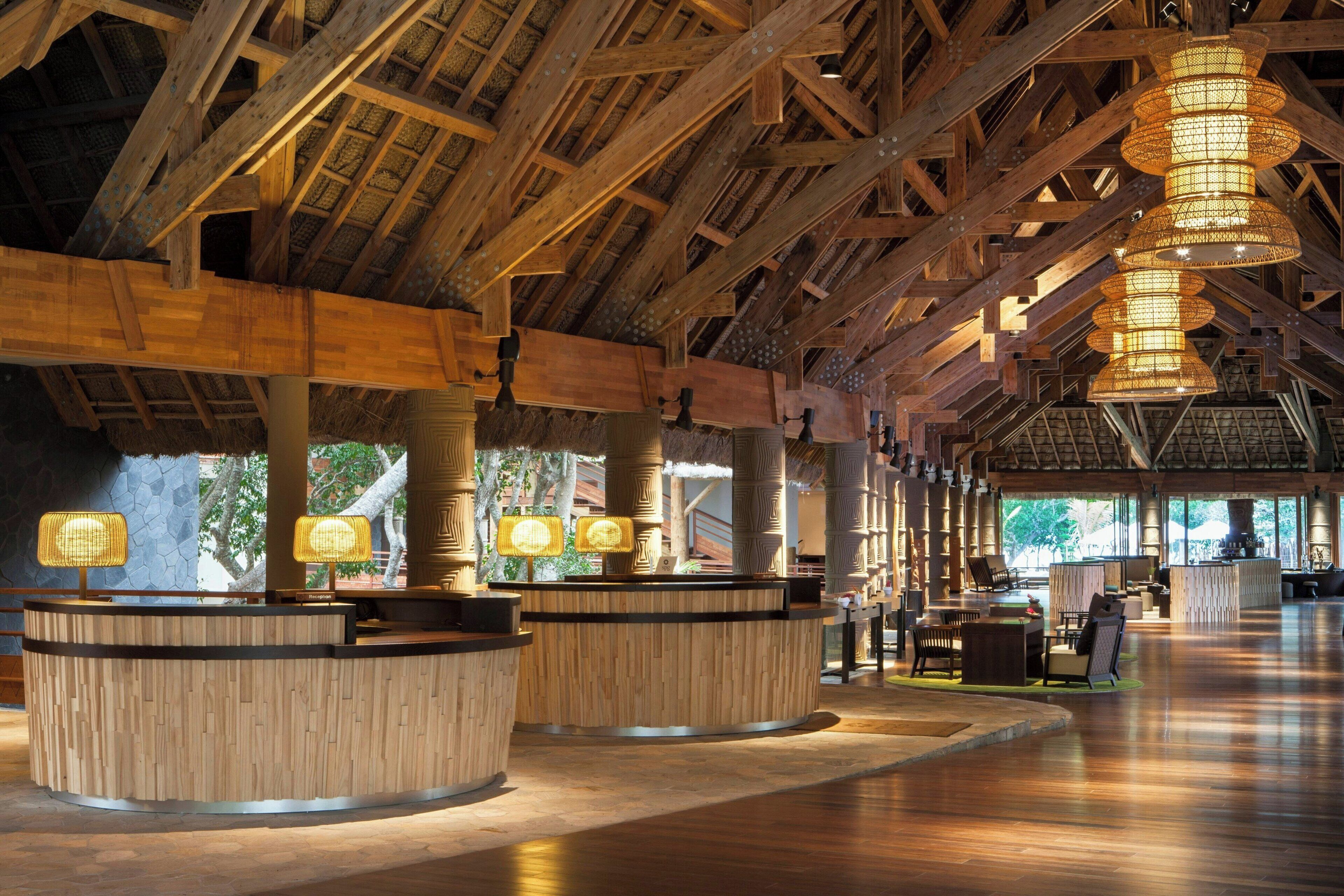 A spacious lobby with wooden reception desks, hanging lamps, and a high ceiling with exposed beams.