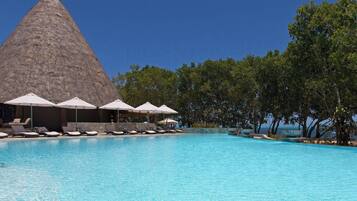A swimming pool with lounge chairs and umbrellas, a thatched-roof structure, and a view of the sea.