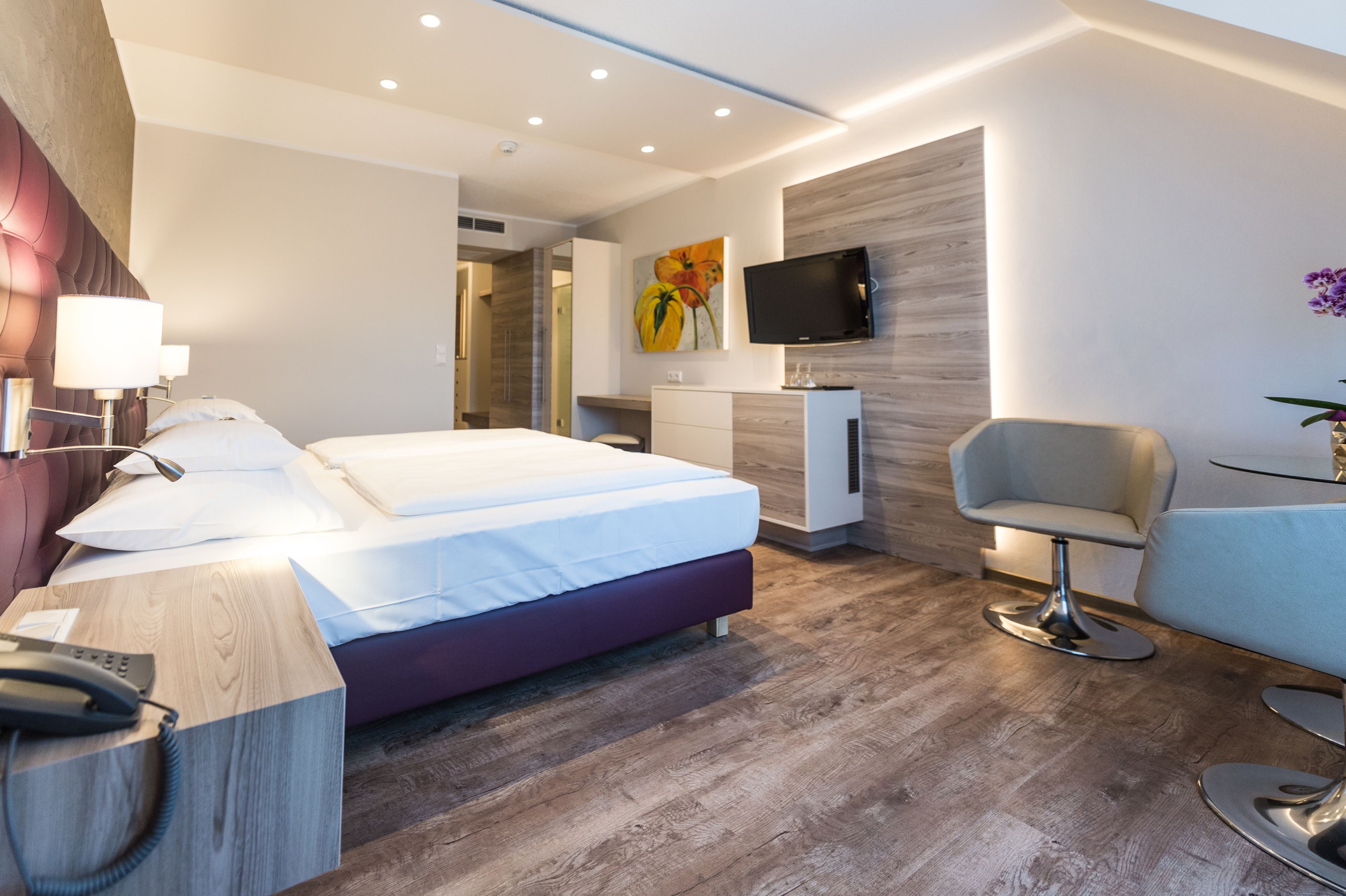 A modern hotel room with a large bed, a desk with a telephone, a TV mounted on the wall, and a seating area with a chair and table.