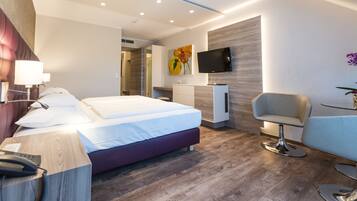 A modern hotel room with a large bed, a desk with a telephone, a TV mounted on the wall, and a seating area with a chair and table.