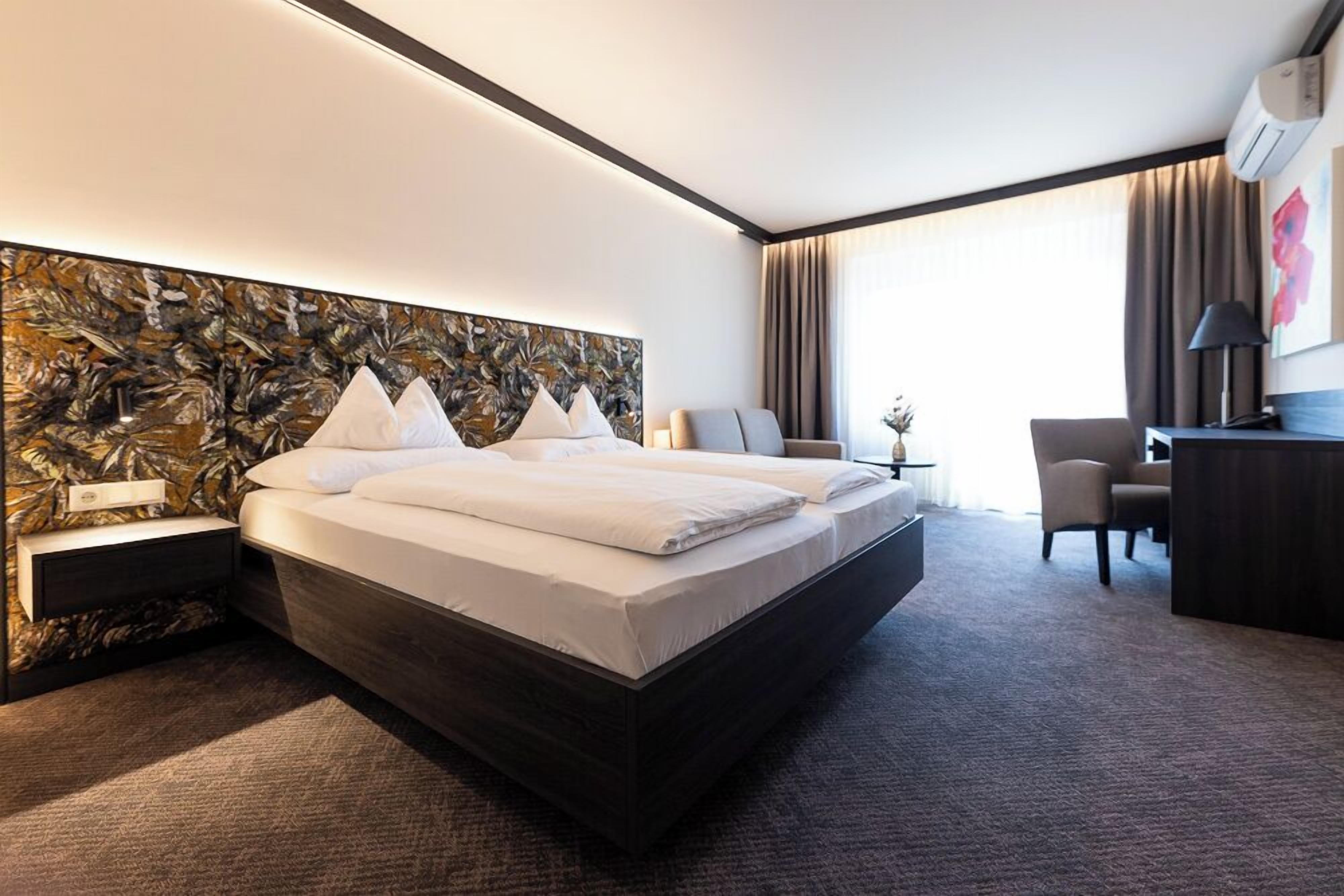A modern hotel room with a large bed, a desk, a chair, and a view of the outdoors.