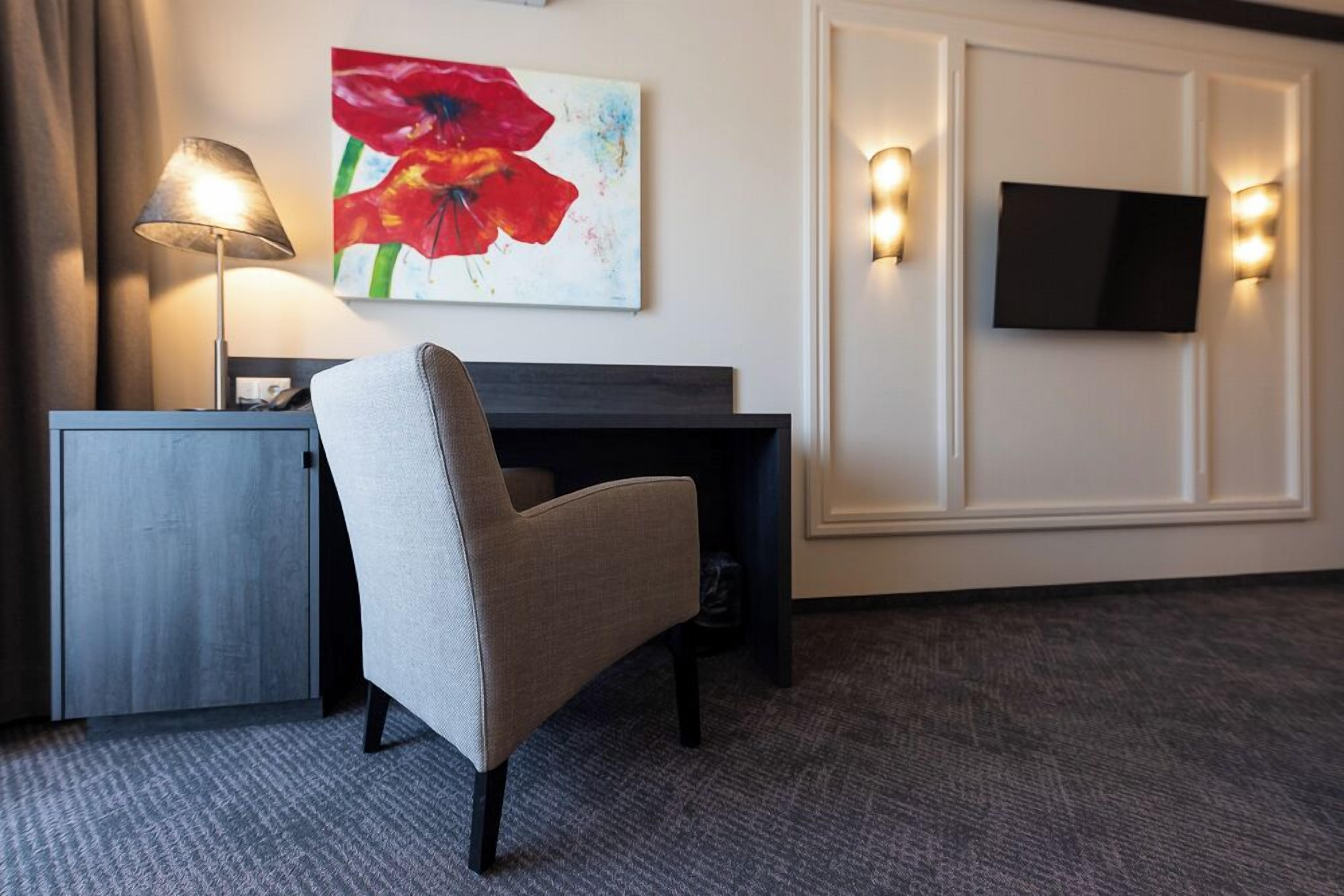 A hotel room with a desk, a chair, a lamp, a TV, and a painting on the wall.