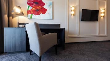 A hotel room with a desk, a chair, a lamp, a TV, and a painting on the wall.