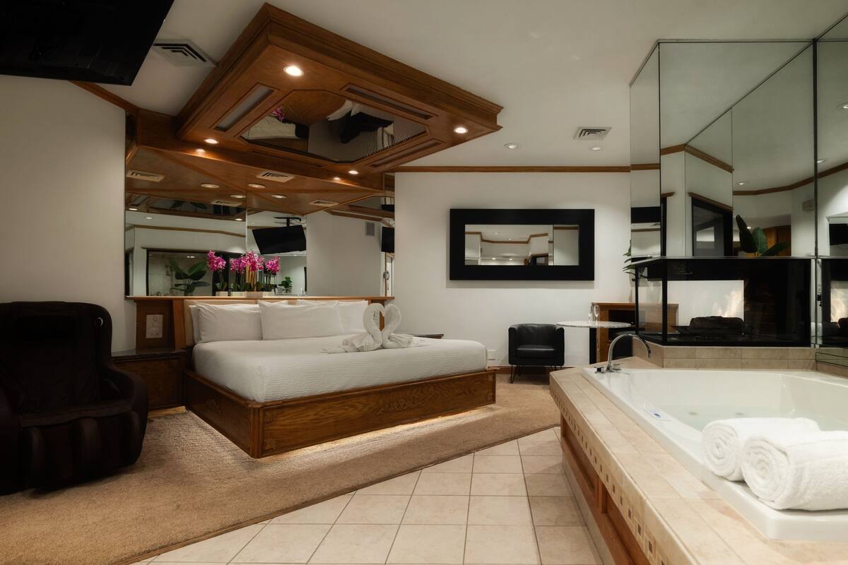 A modern bedroom with a large bed, a separate sitting area, a glass-enclosed bathroom, and a large mirror.