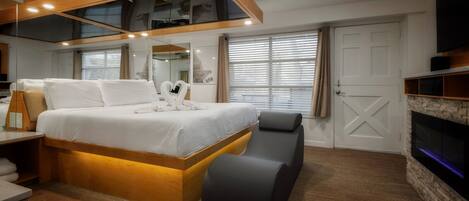 A modern hotel room with a large bed, a bench, a flat-screen TV, and a fireplace.