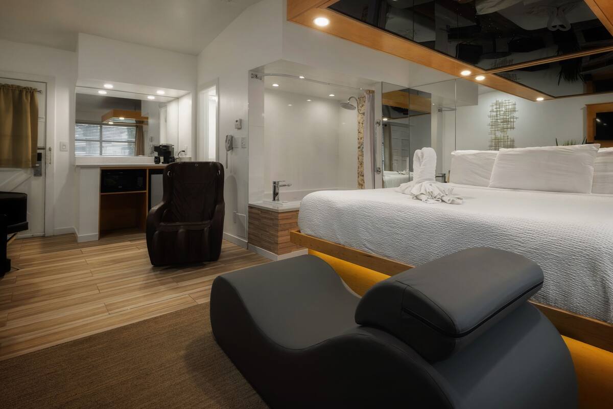A modern hotel room with a large bed, a massage chair, a small desk, and a shower area.