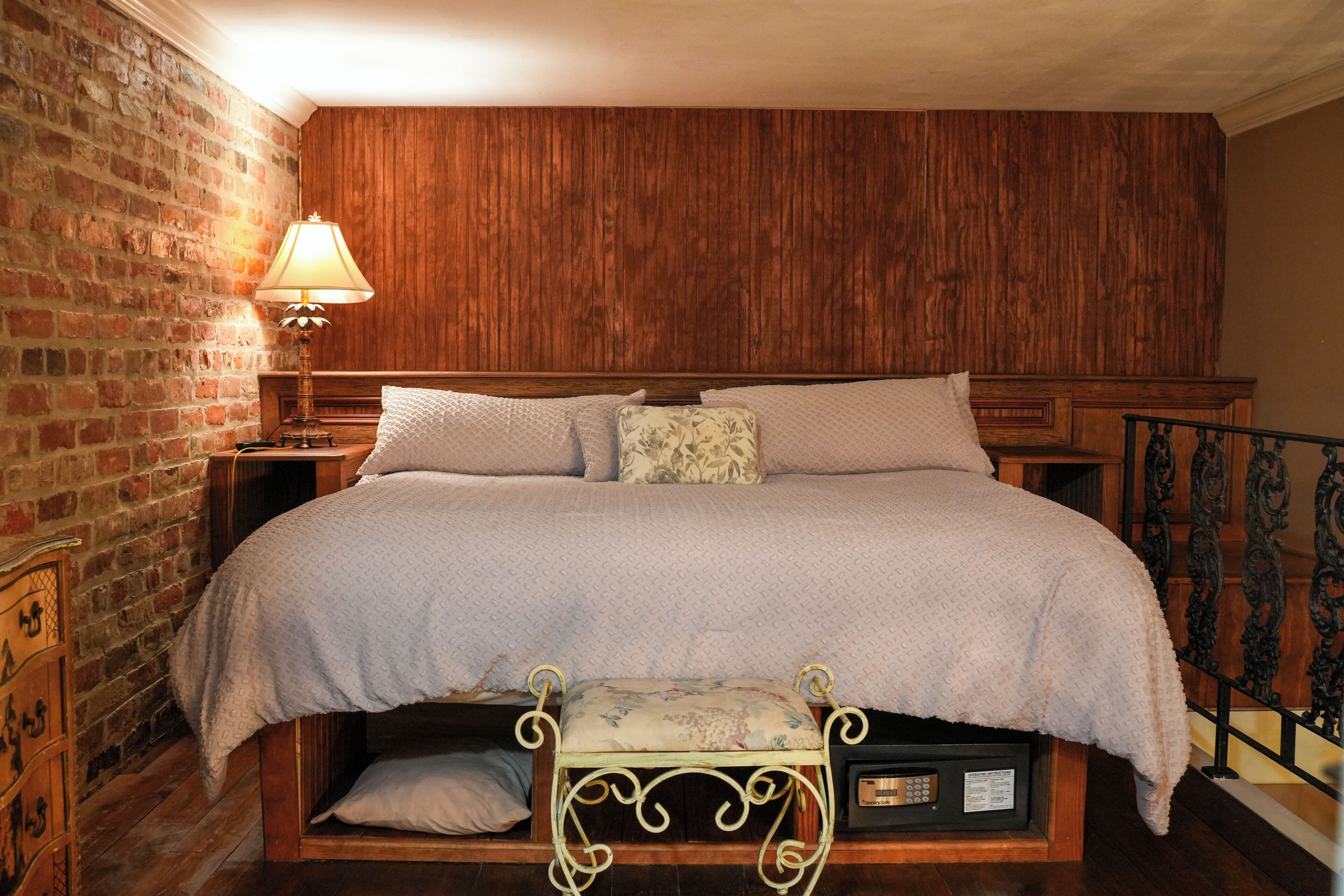 A bed with a wooden headboard, a bench with a cushion, a nightstand with a lamp, and a brick wall.
