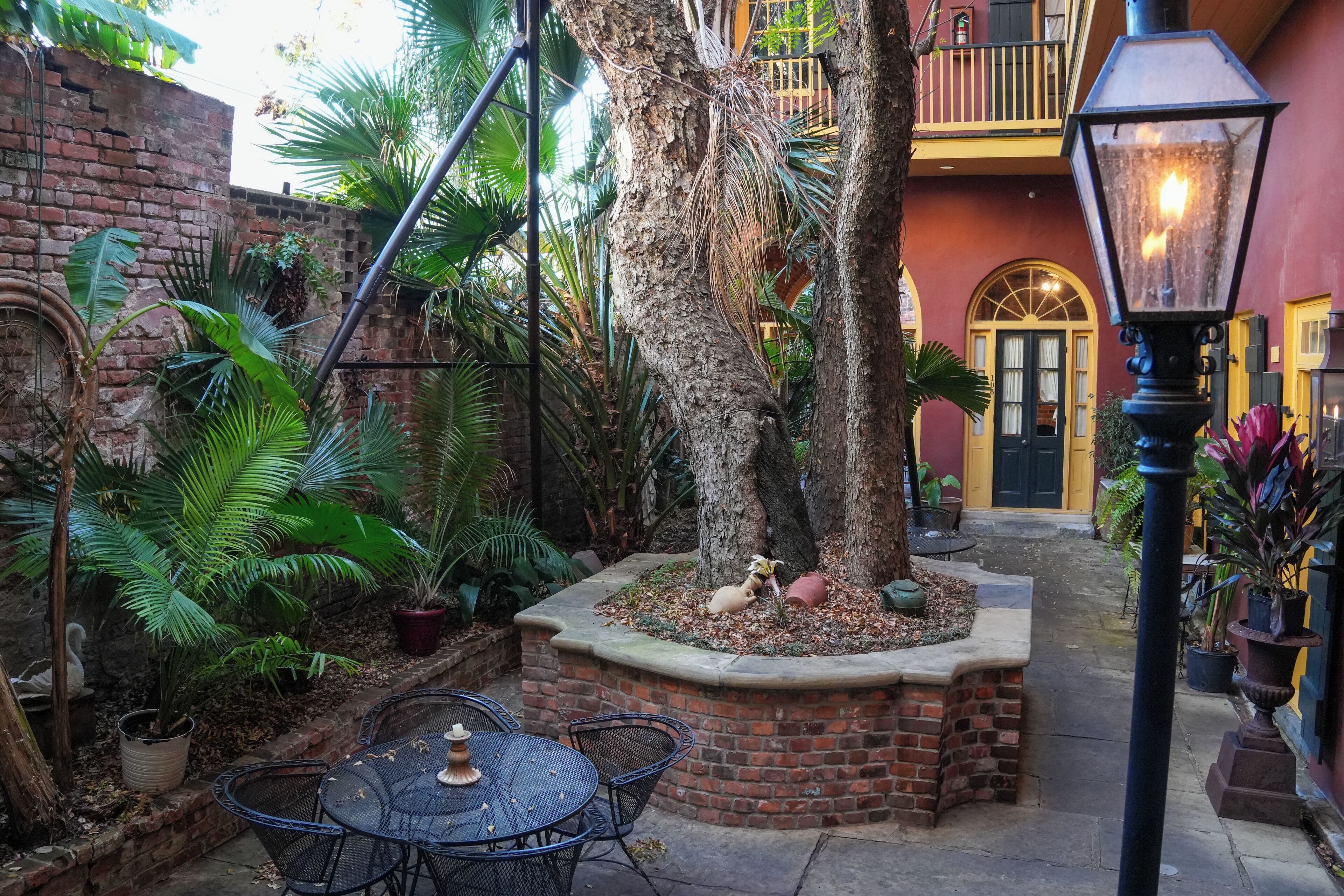 A courtyard with a brick wall, palm trees, a table with chairs, and a lantern.