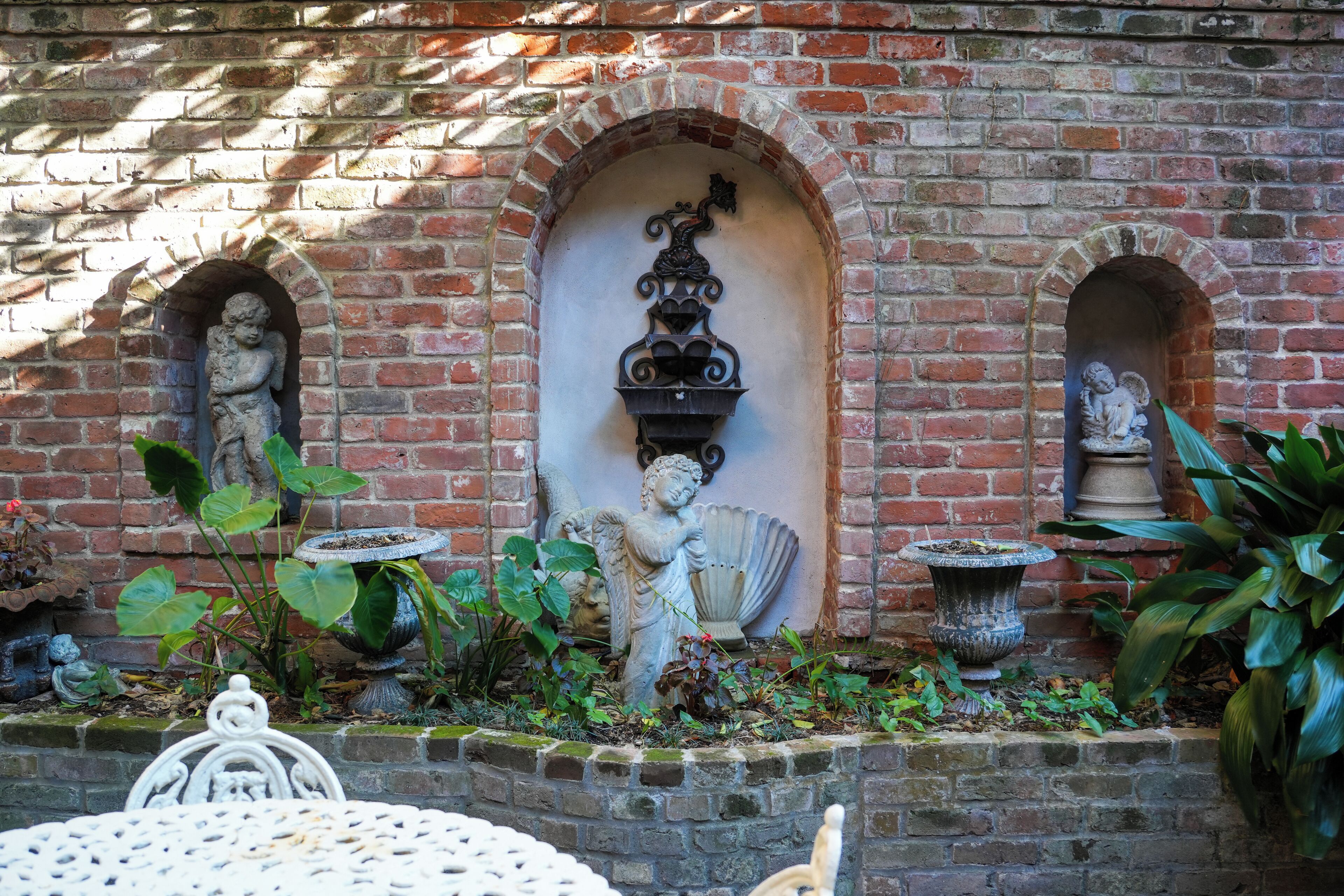 A brick wall with three arched niches, each containing a statue. There are potted plants and a bird bath in the foreground.