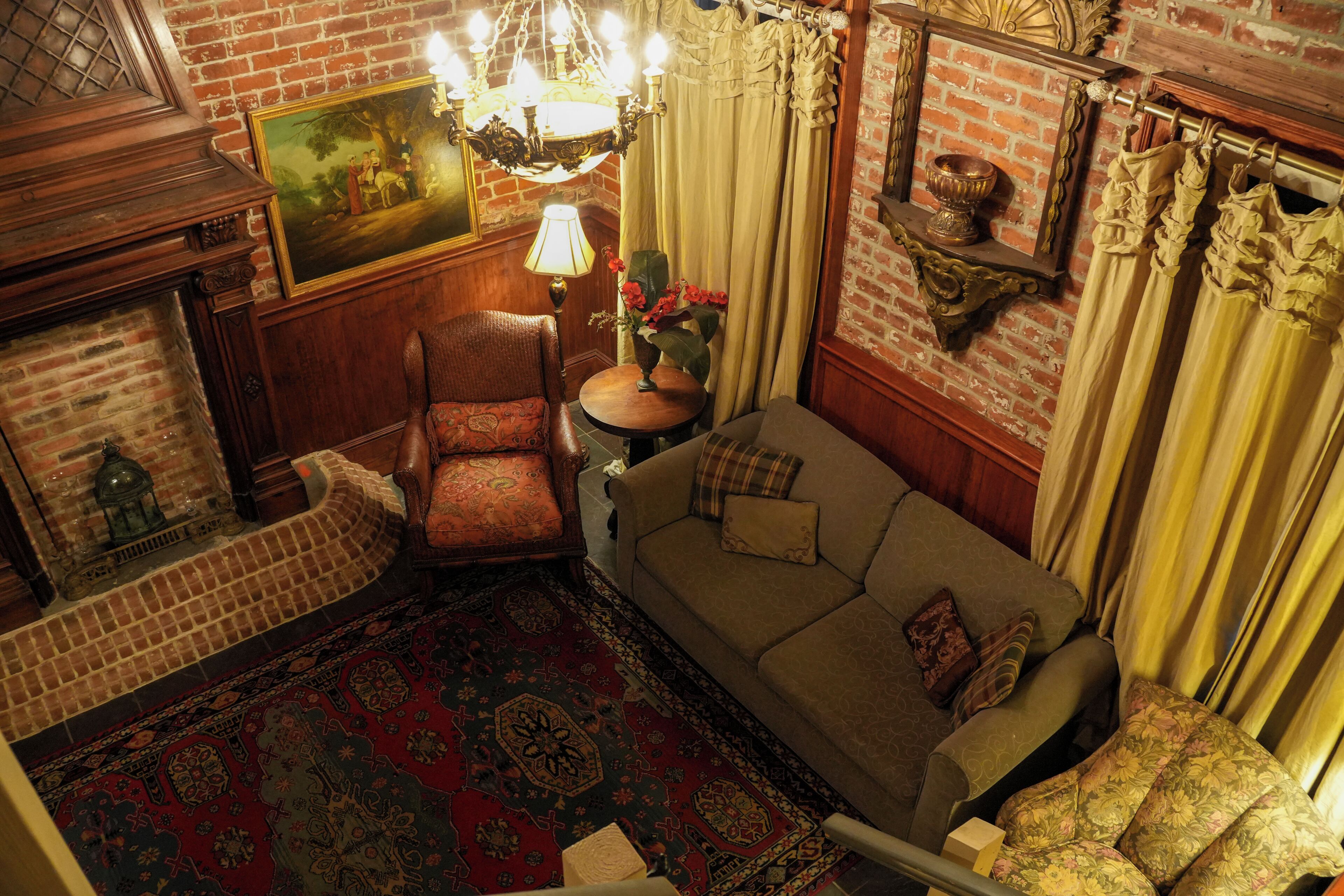 A room with a brick wall, a wooden sofa, a patterned rug, a wooden chair, a table with a vase, and a chandelier.