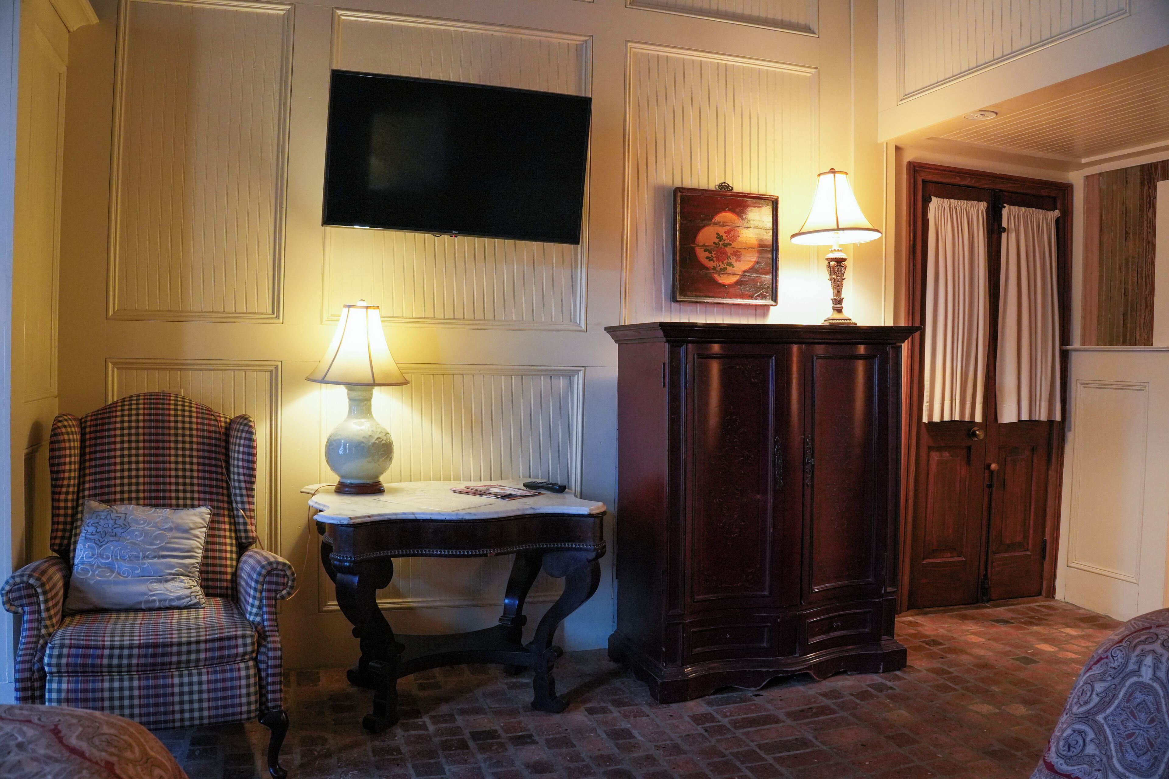 A room with a plaid armchair, a small table with a lamp, a wooden cabinet, and a television mounted on the wall.