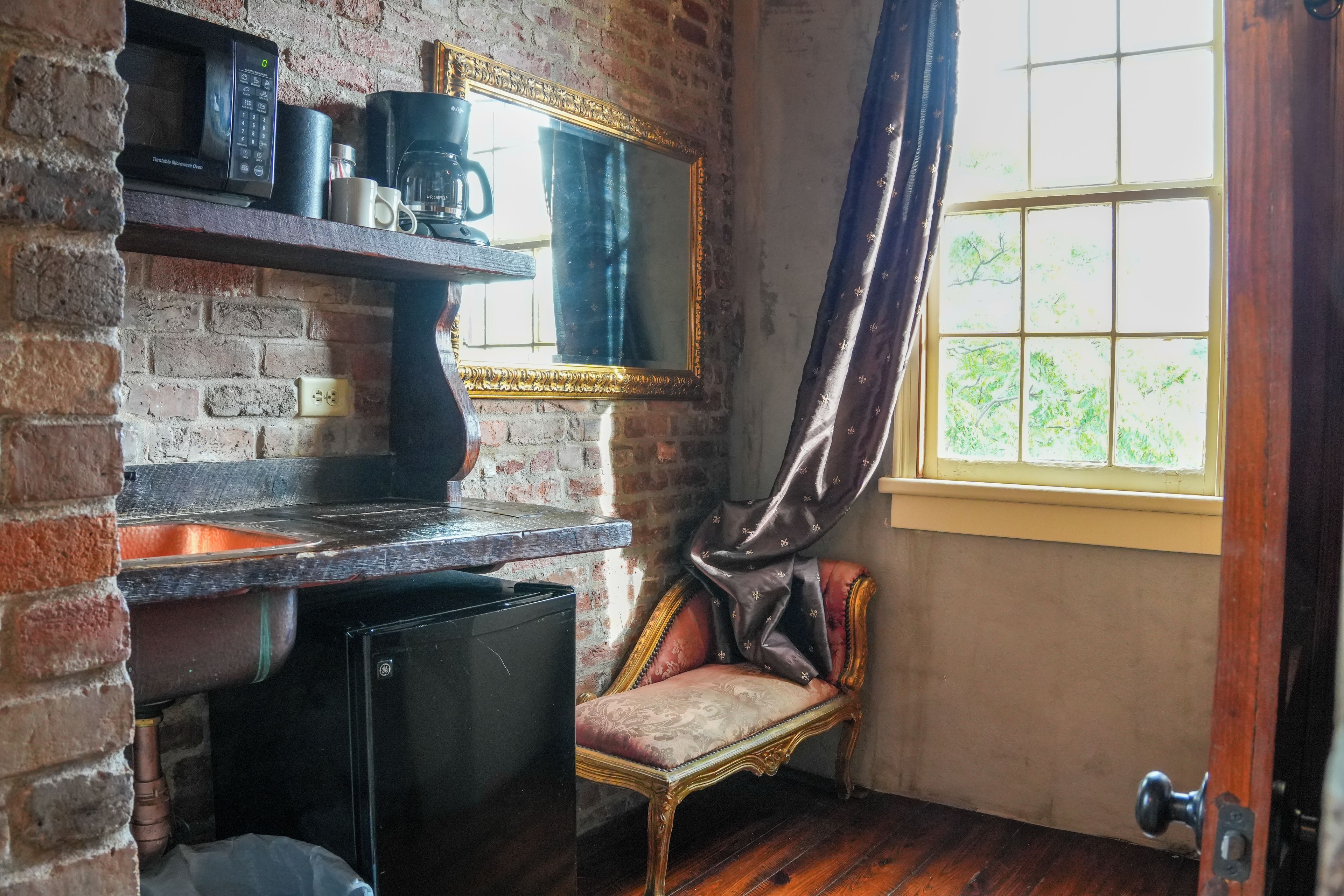 A room with a brick wall, a small black refrigerator, a coffee maker, a mirror, and a window with curtains.
