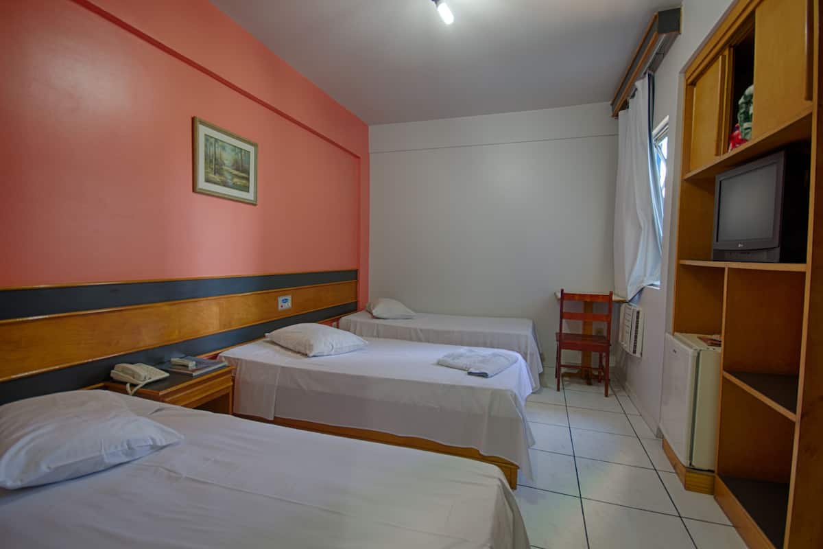 Habitación de hotel con tres camas individuales, cabecera de madera, una mesita con teléfono, una silla, televisor sobre un estante de madera y un cuadro enmarcado en la pared.