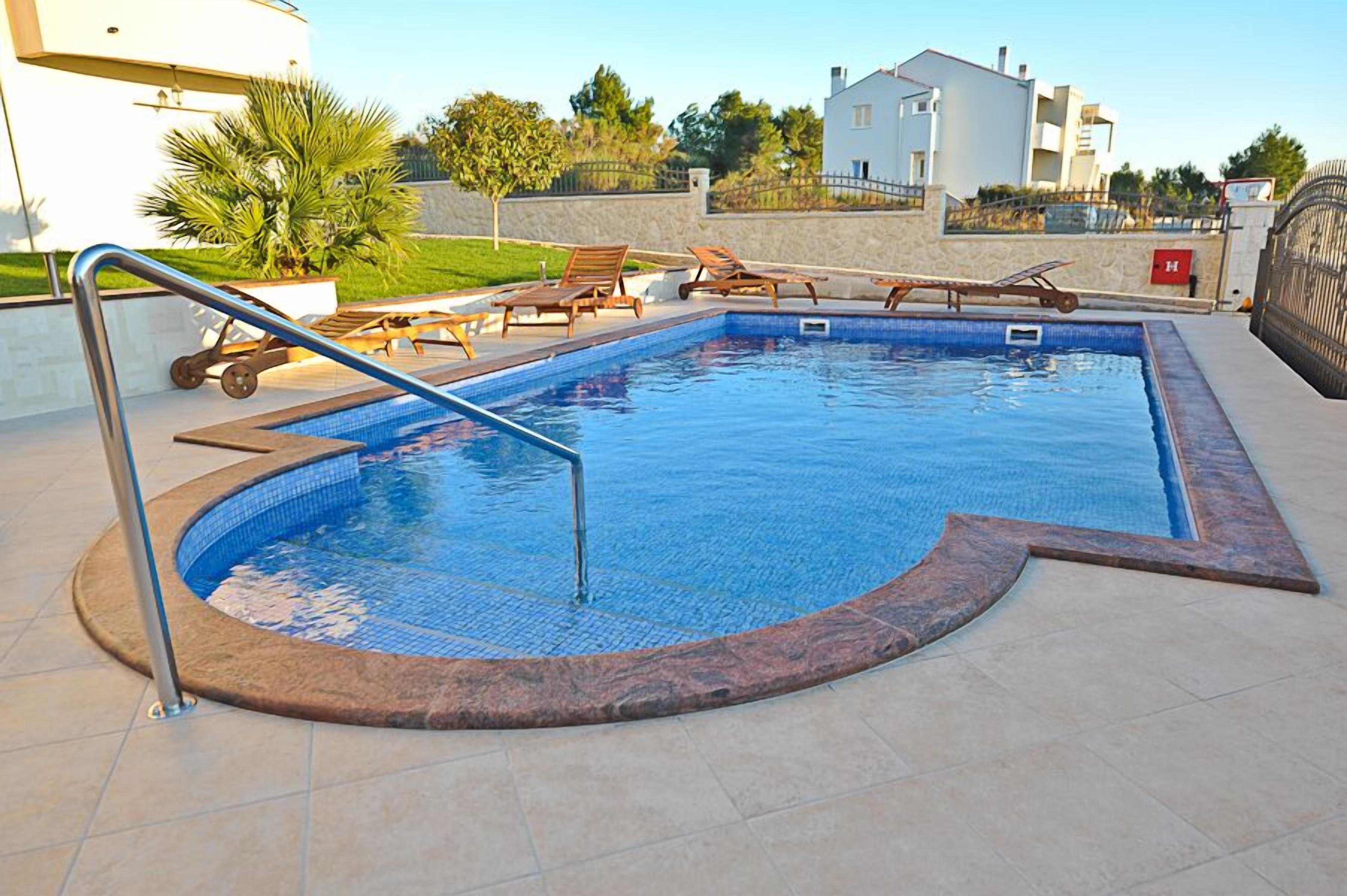 Ein rechteckiger Swimmingpool mit gefliestem Rand, umgeben von Liegestühlen und einem Metall-Geländer.