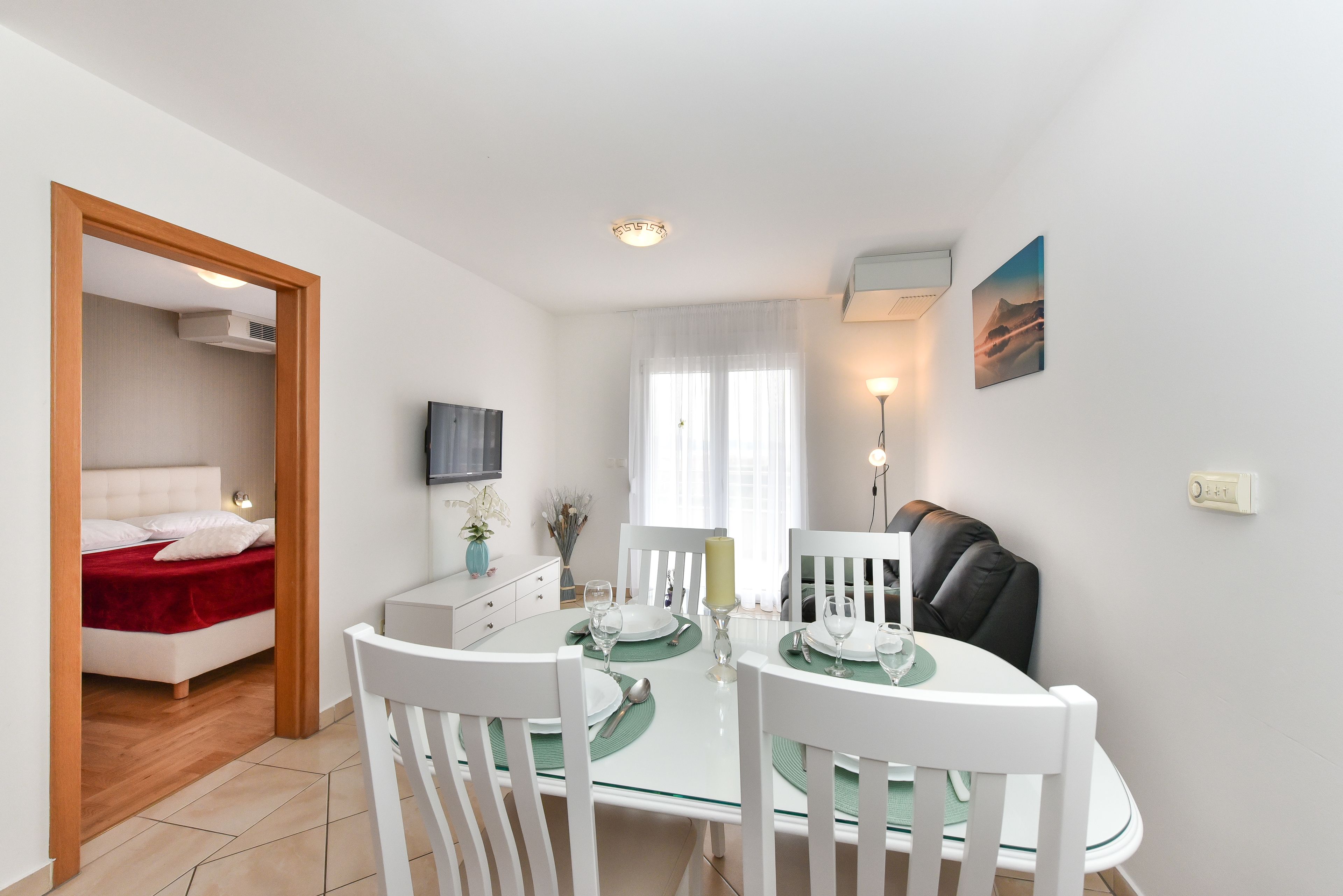 Un espace repas moderne avec une table en verre, des chaises blanches et un lit rouge en arrière-plan.