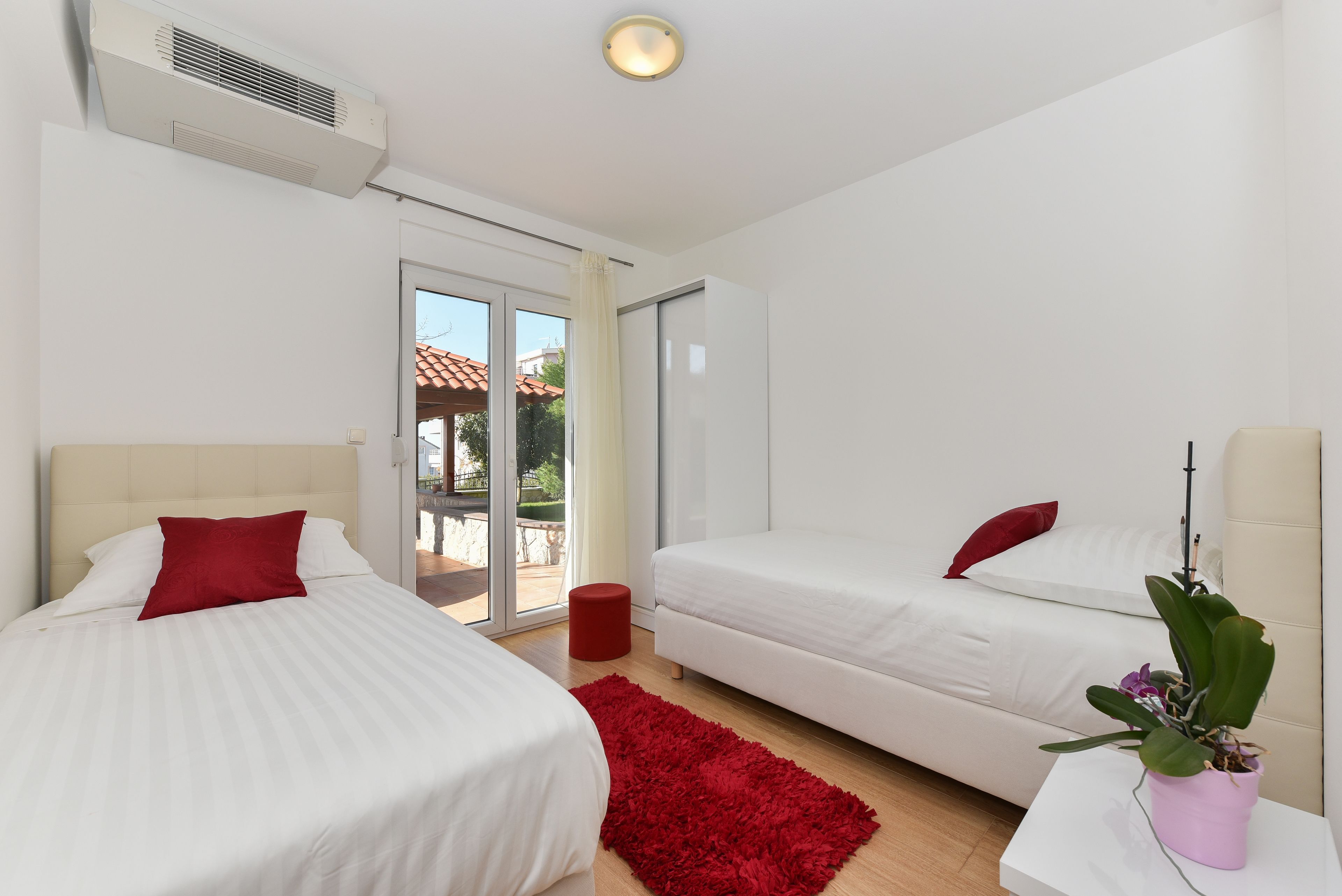Una habitación de hotel moderna con dos camas, una alfombra roja, una planta en maceta y un ventanal corredero que da a un espacio exterior.
