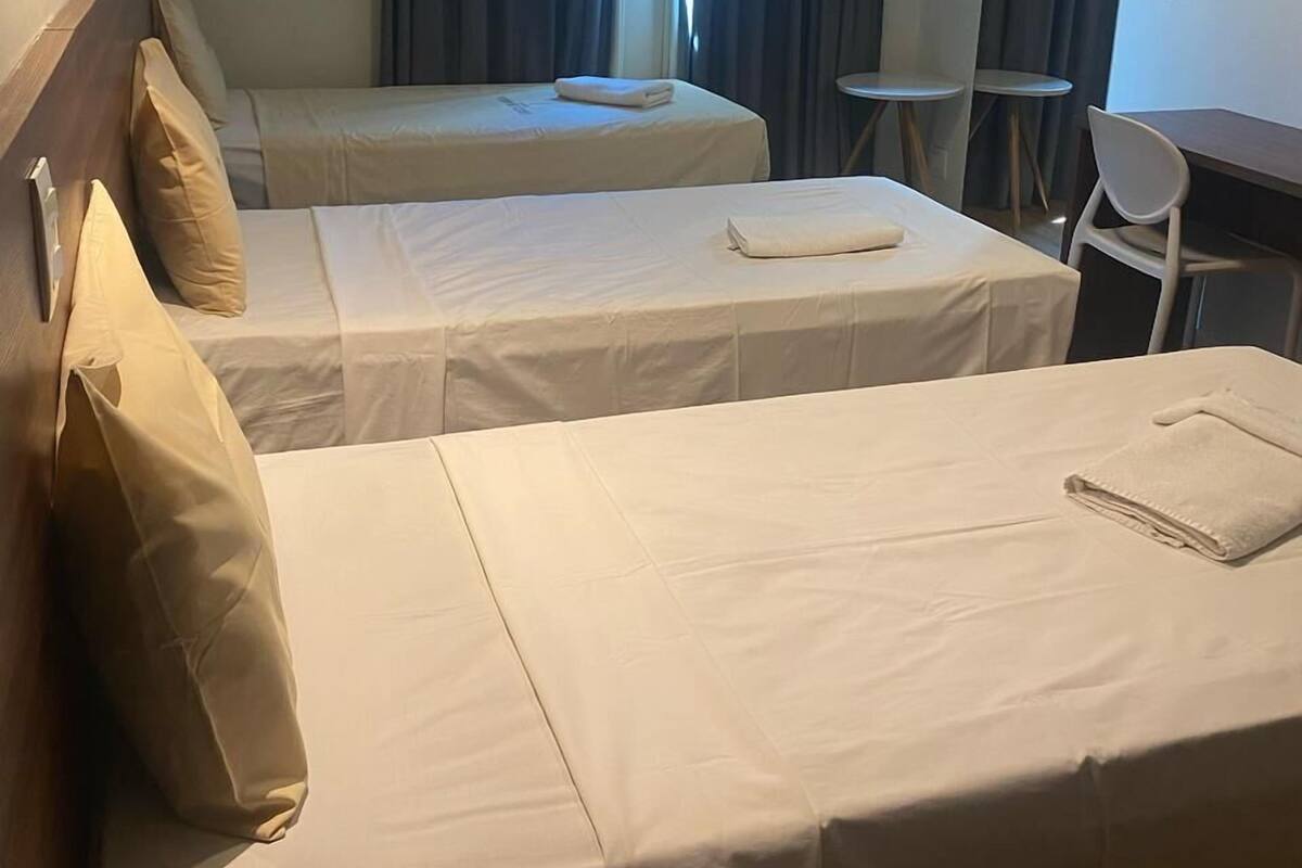 A hotel room with two single beds, a small table, a chair, and a window with curtains.