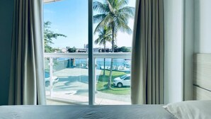 A hotel room with a large window, a bed, and a view of a parking area with cars and palm trees. - Ibiza Barra Hotel (Rio de Janeiro)