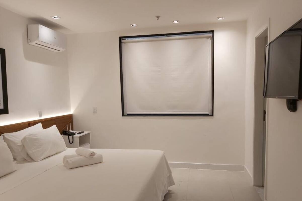 A hotel room with a bed, a bedside table with a phone, a wall-mounted air conditioner, and a large window with blinds.