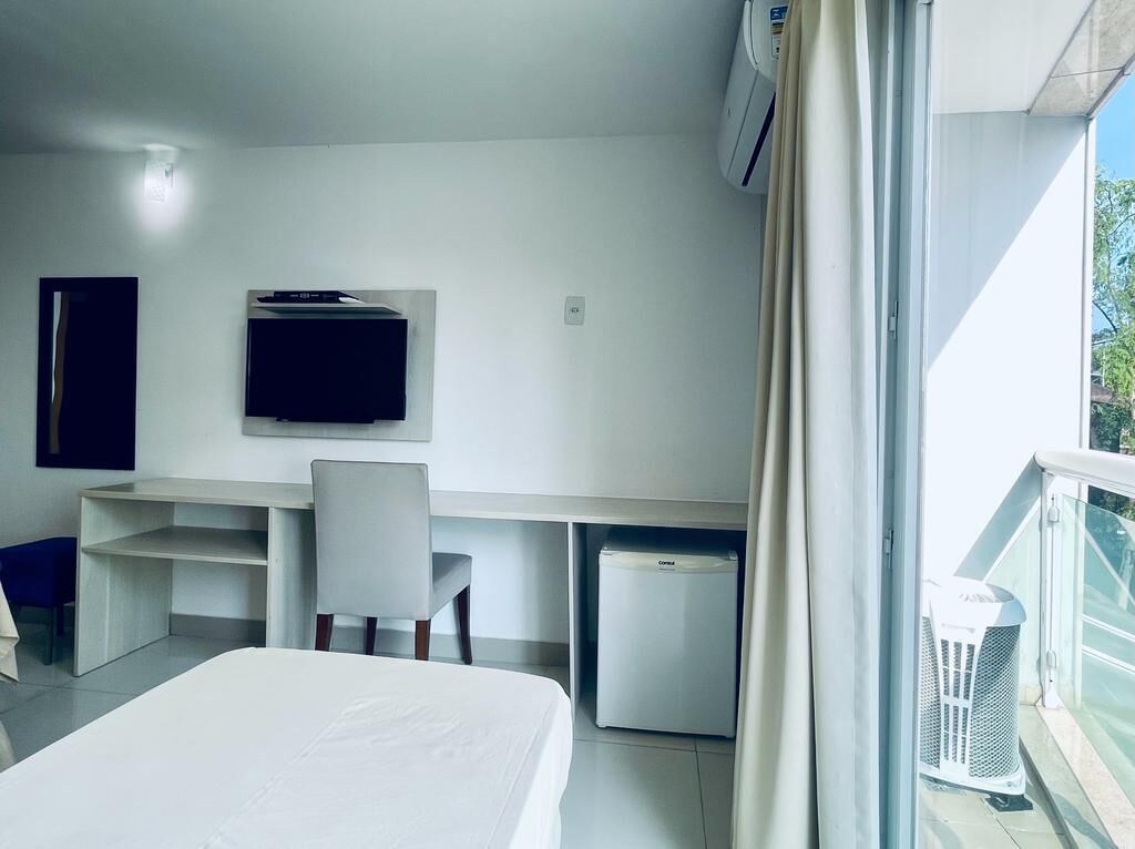 A hotel room with a bed, a desk with a chair, a television, a small refrigerator, and a balcony with a railing.
