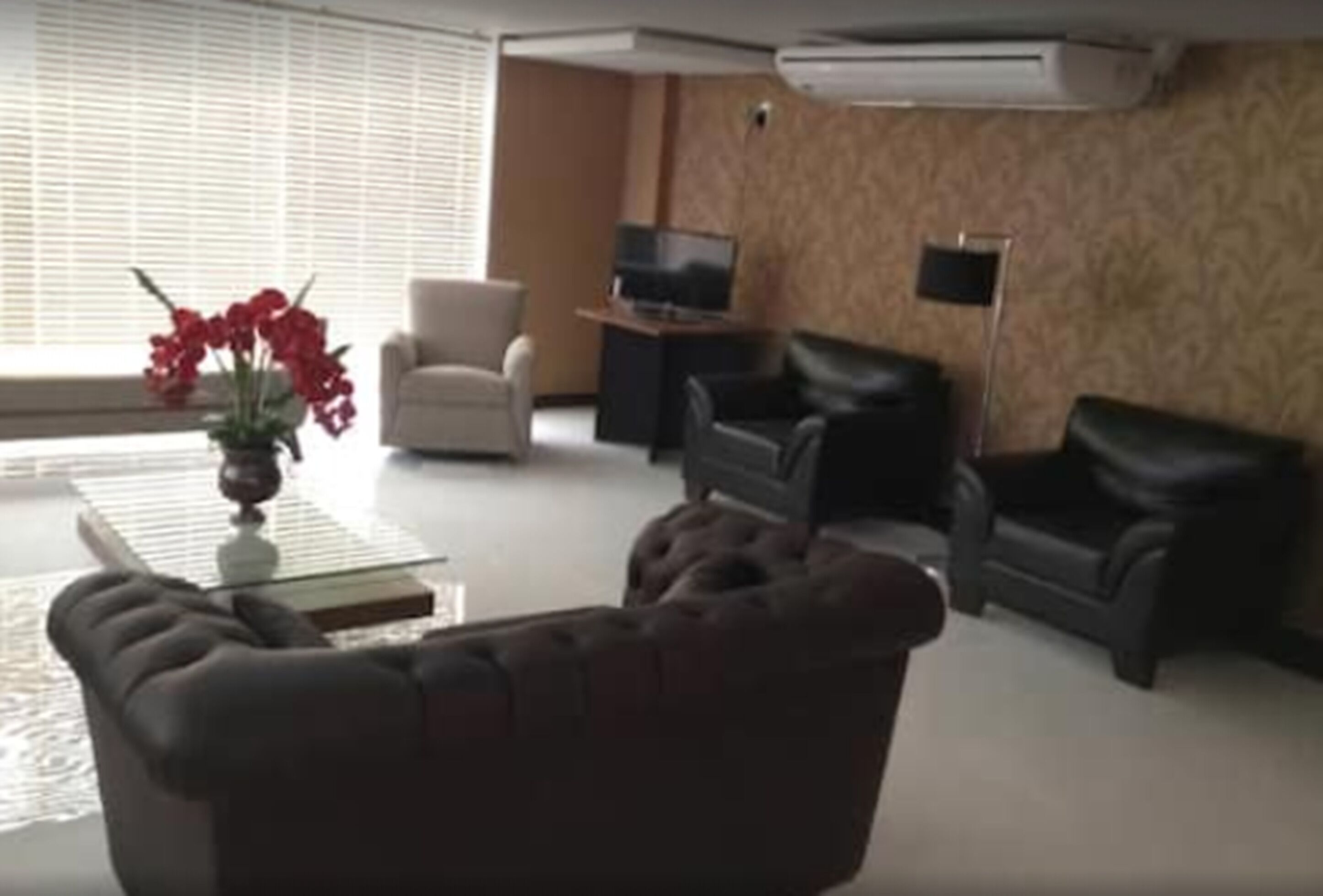 Un salón con un sofá marrón, un sillón blanco, un sofá negro, un televisor sobre un mueble bajo y un arreglo floral rojo.