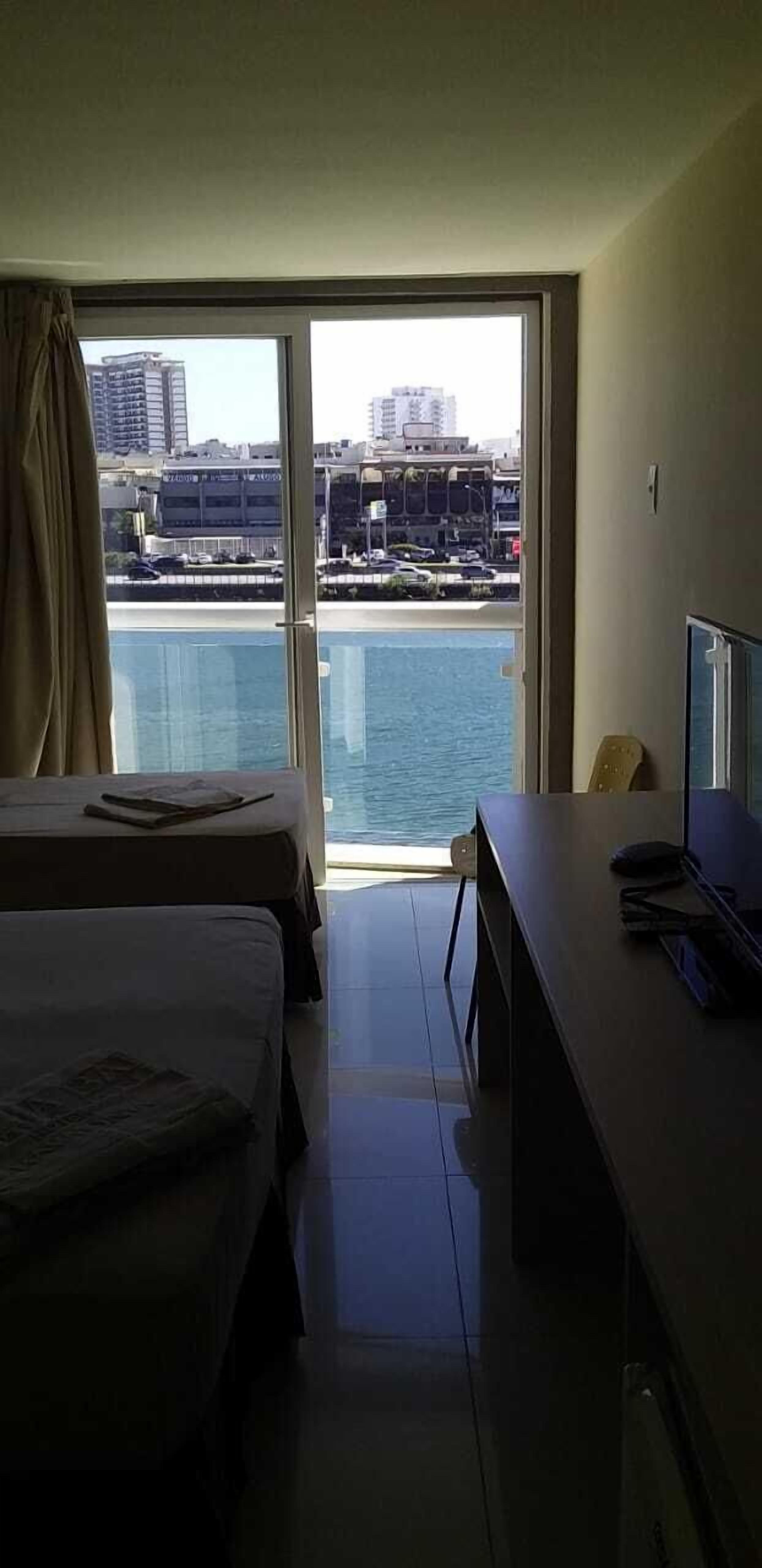A hotel room with a large window, a bed, a desk, and a view of a body of water and buildings.