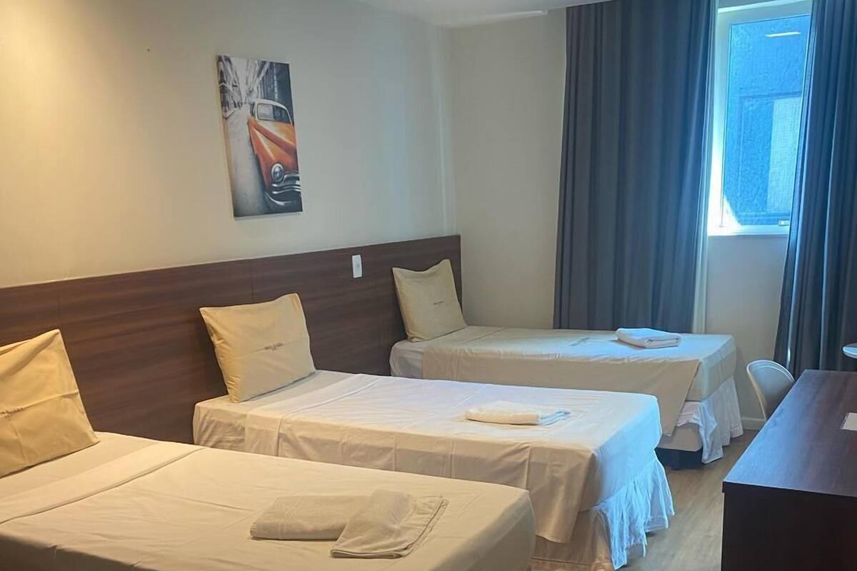 A hotel room with two single beds, a wooden headboard, a painting on the wall, and a window with curtains.
