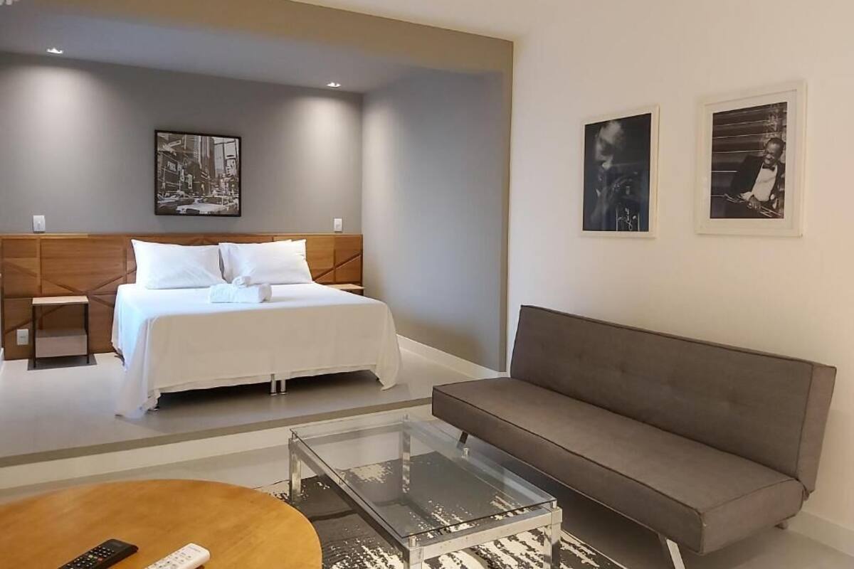 A modern hotel room with a bed, a gray sofa, a glass coffee table, and artwork on the walls.