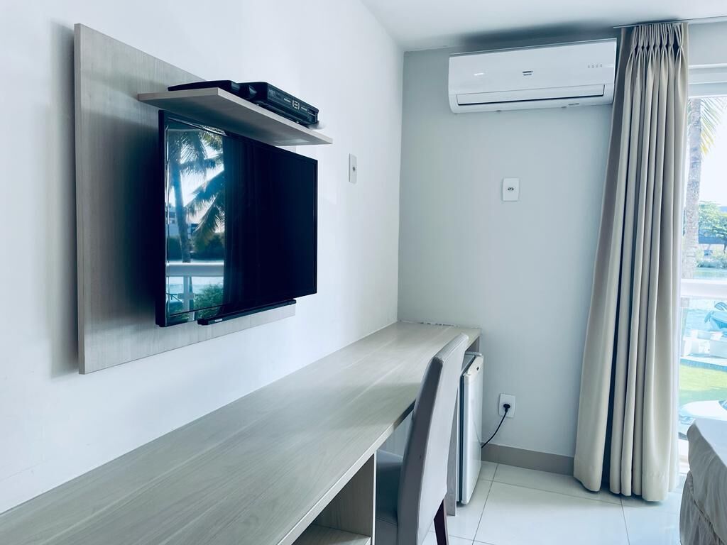 A modern room with a flat-screen TV mounted on the wall, a small desk with a chair, an air conditioning unit, and a view of palm trees through the window.