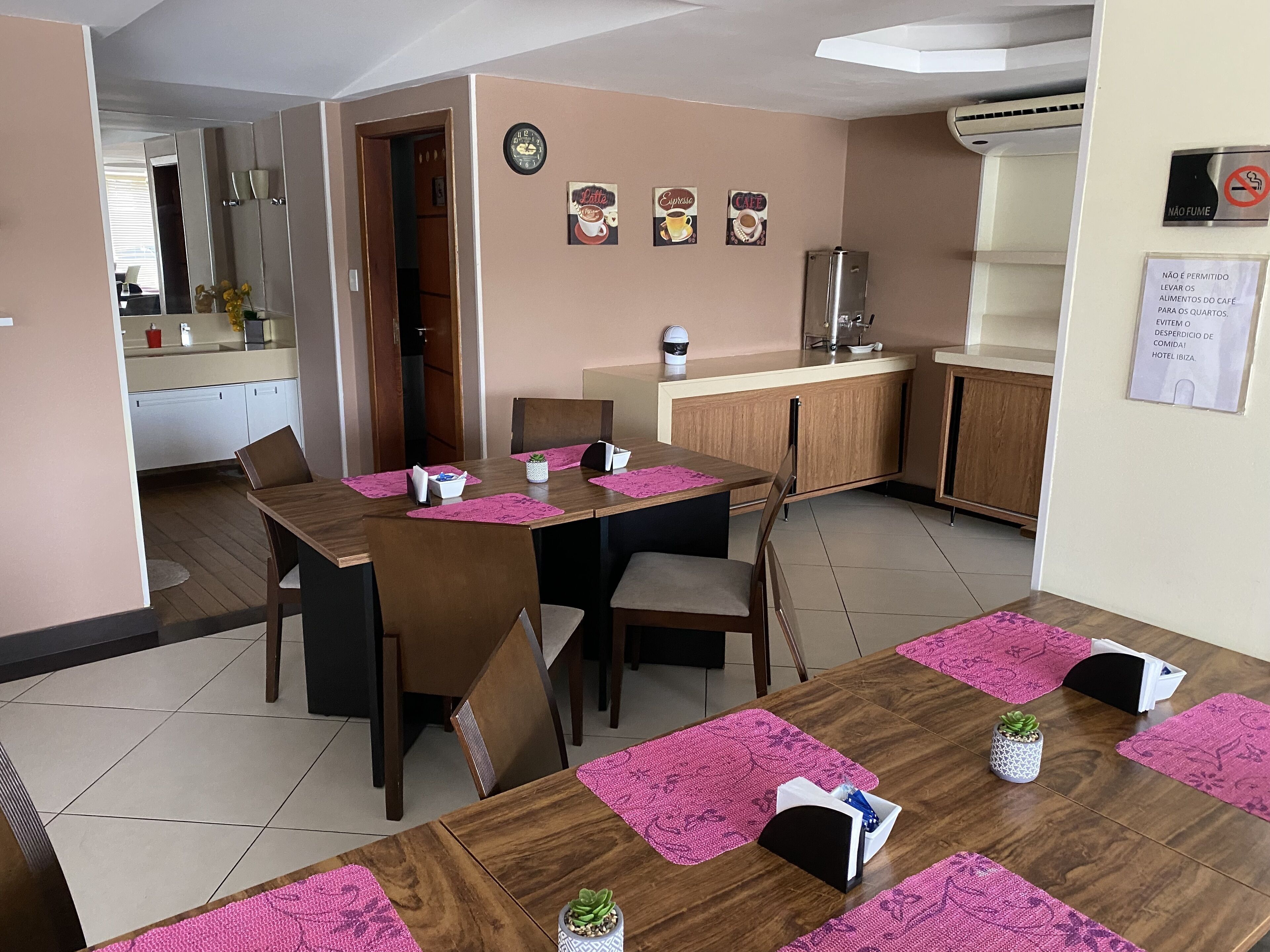 A dining area with wooden tables and chairs, pink placemats, and a kitchenette in the background.