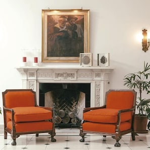 A room with a fireplace, two armchairs, a framed painting above the fireplace, and a potted plant. - Solar De La Plaza (Salta)