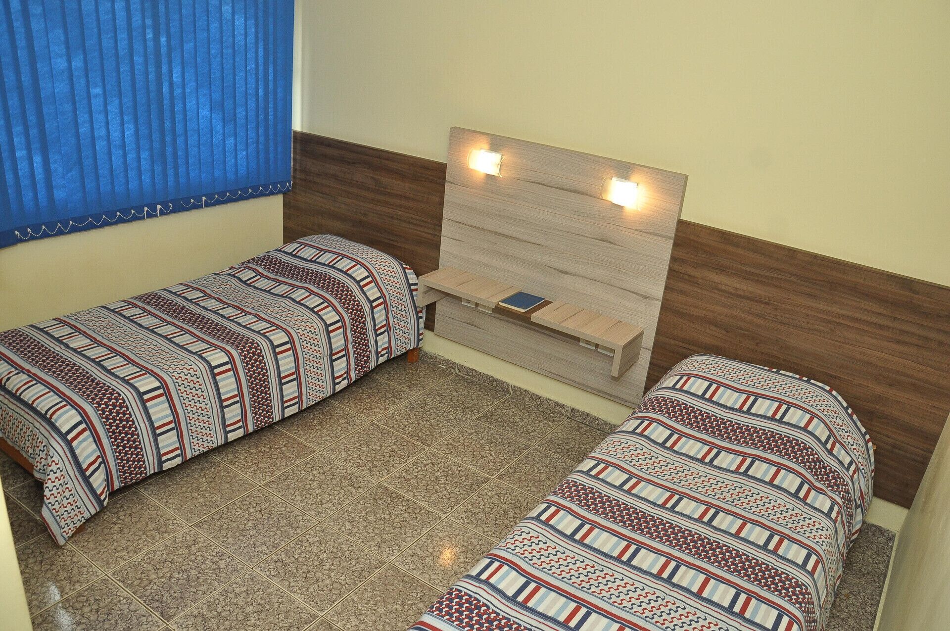 Una habitación pequeña con dos camas individuales, un cabecero de madera y una ventana con cortinas azules.