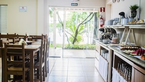 Zona ristoro di un hotel con tavoli e sedie in legno, un banco buffet con diversi alimenti e un cartello che indica l'uscita.