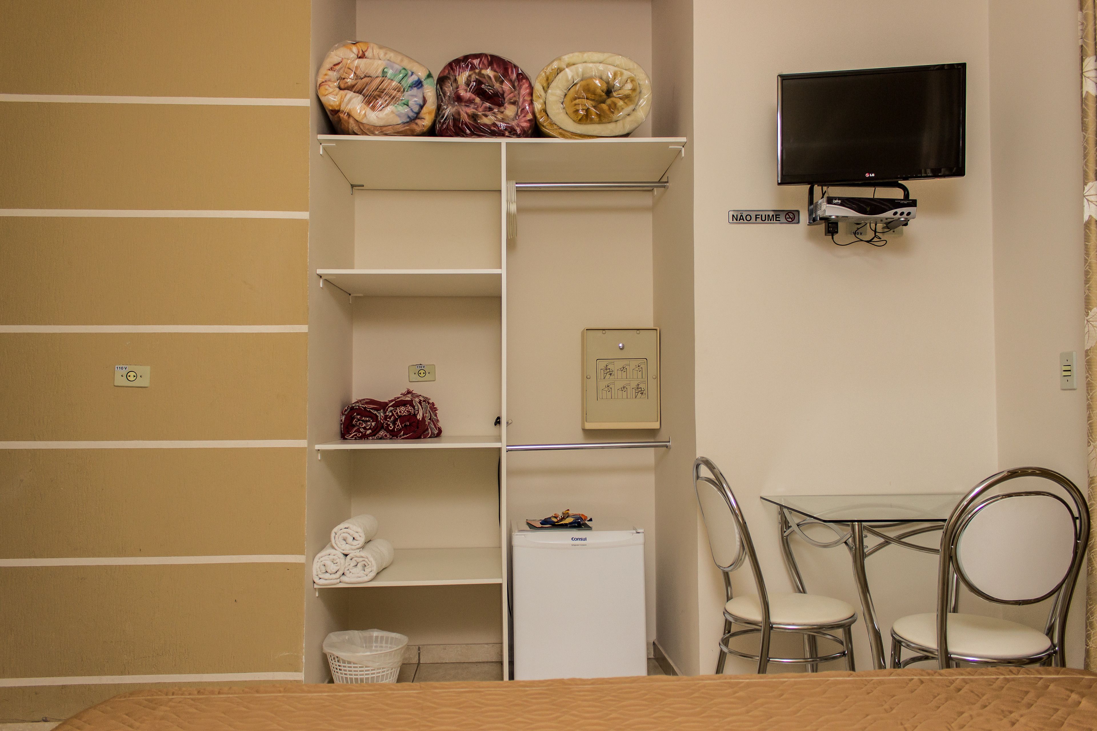 Une pièce de taille modeste comprenant un lit, une petite table avec des chaises, un téléviseur fixé au mur et des étagères contenant divers objets.