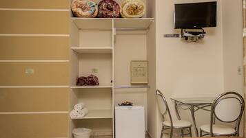 Une pièce de taille modeste comprenant un lit, une petite table avec des chaises, un téléviseur fixé au mur et des étagères contenant divers objets.